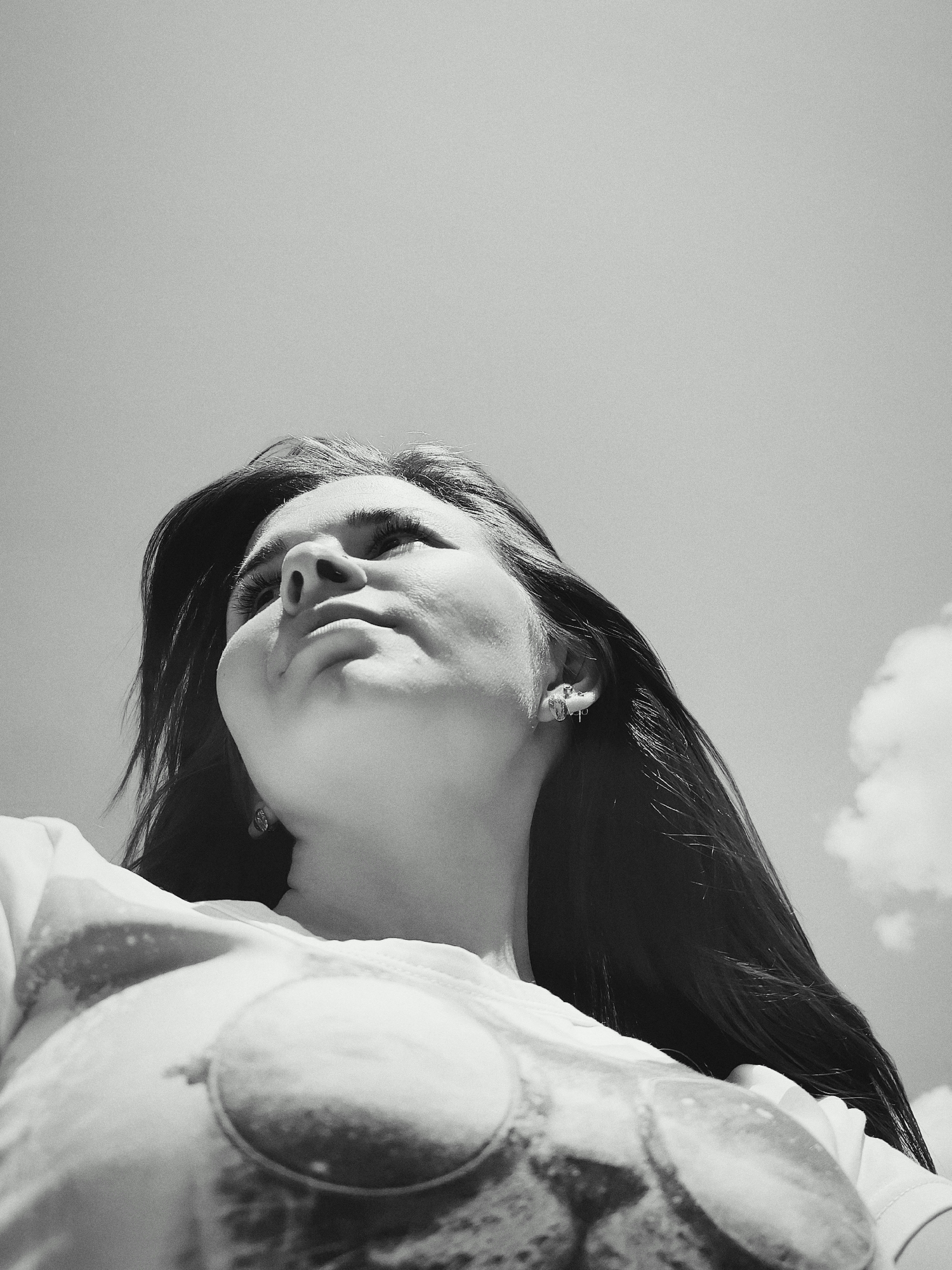 A black and white, low-angle photograph depicts a woman looking upward. The sky is visible, with a cloud in the lower right corner. The woman has long hair and is wearing earrings. The focus and perspective make the details of her shirt less clear, but a circular pattern is visible. There is no indication of a specific location within Фоки, Russia, or any identifying details beyond the person, sky, and cloud.