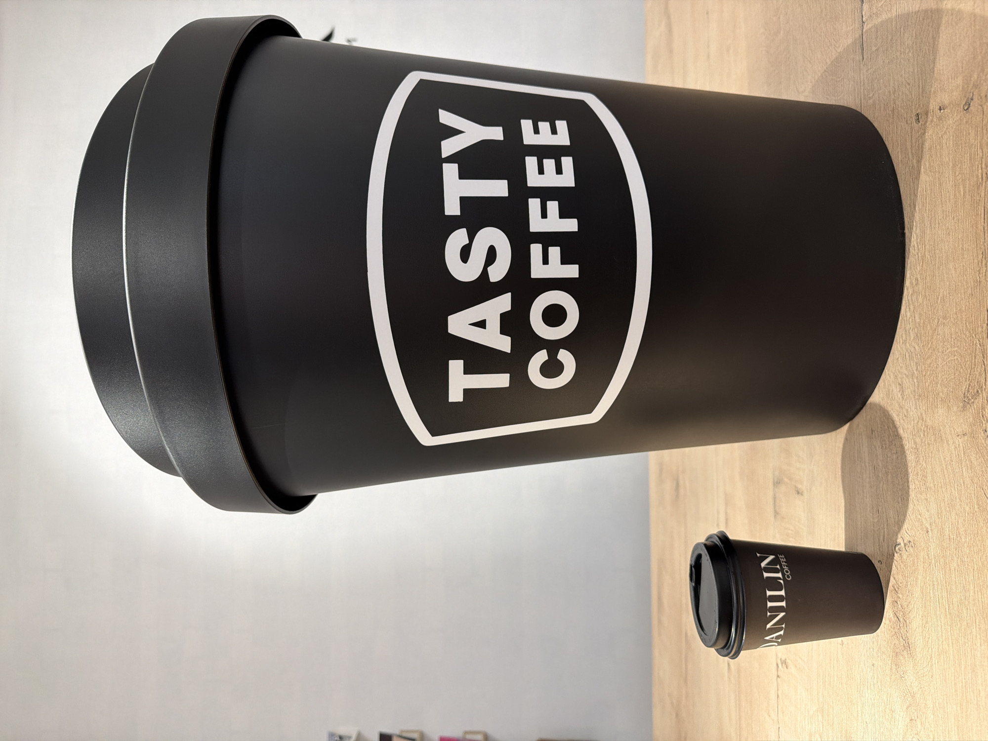 The image features an oversized black coffee cup with a white logo that reads "TASTY COFFEE." Next to it is a smaller, regular-sized brown coffee cup with a black lid that has the name "VANILIN COFFEE" on it. Both items are placed on a wood-grain surface. The background includes a white wall, suggesting an indoor setting. This image was taken in Chaikovsky, Russia.