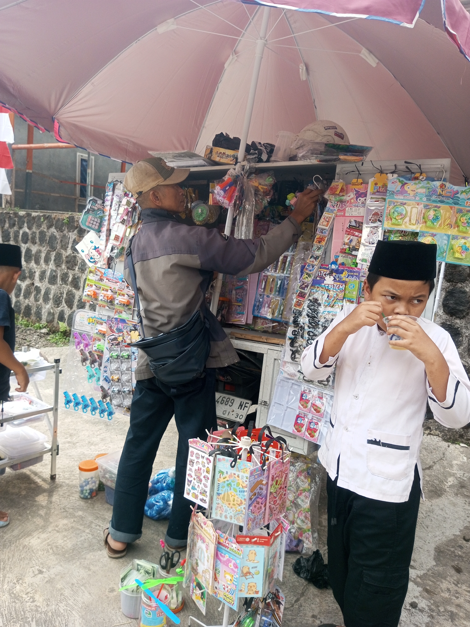 A young boy wearing a white Koko shirt and black peci, is drinking from a clear plastic cup while standing in front of a stall selling toys. The stall is covered by a large pink umbrella. The boy's right hand holds the cup to his lips, and his left hand rests on a straw. The stall displays various toys, including colorful stickers, small plastic toys, and novelty items. A man in a gray jacket, a beige cap, and dark pants is seen in the background, appearing to interact with the stall's merchandise. The scene appears to be outdoors, with a stone wall and some vegetation visible in the background. The ground is paved, and a motorcycle license plate that reads "4689 NF" is partially visible. The location is identified as Krasak, Indonesia.