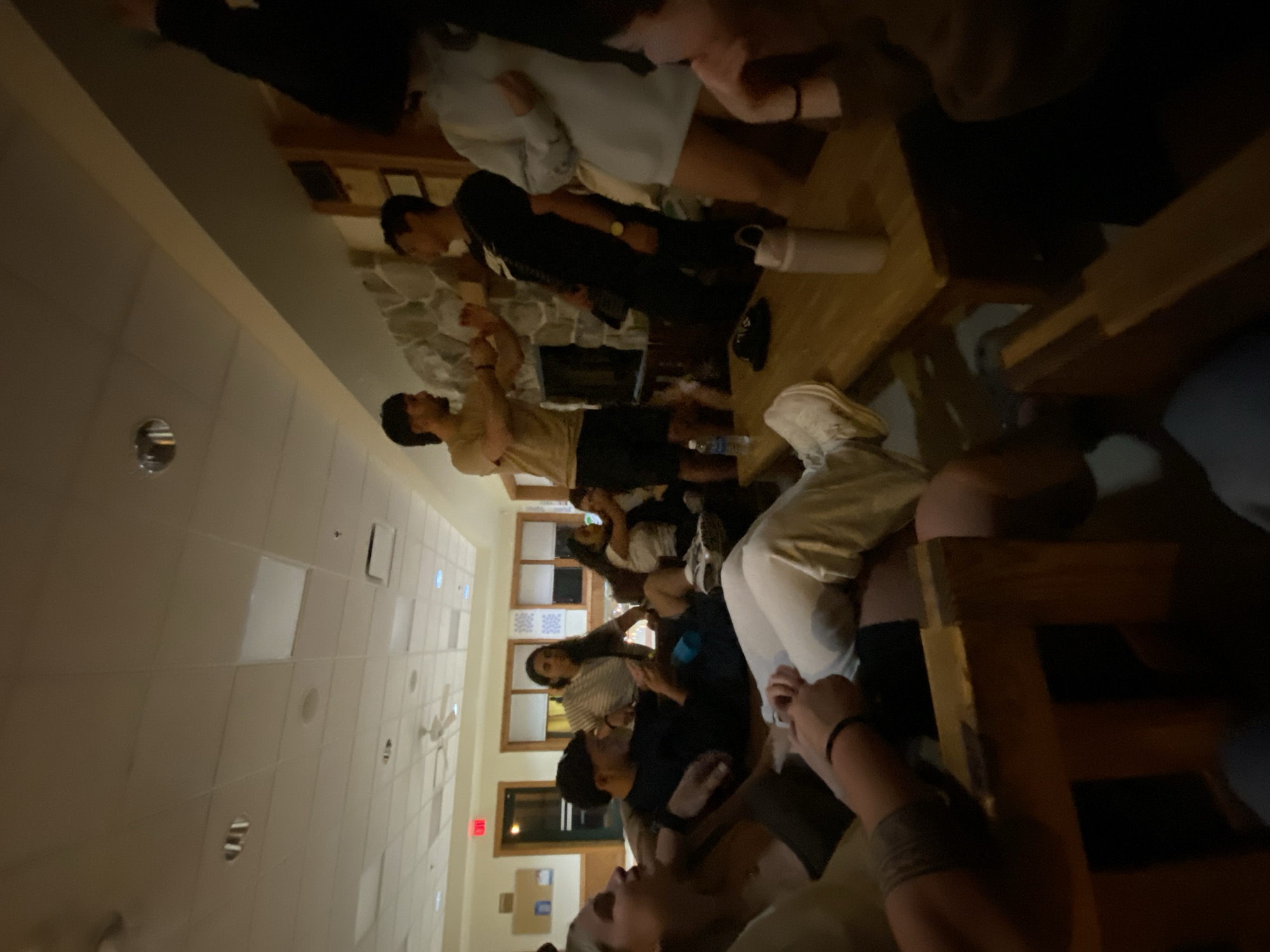 The image depicts a dimly lit, indoor gathering of approximately ten young adults in a common room or lounge area, possibly in a hotel or dormitory, in Washington Township, USA. Several individuals are seated on couches and chairs, while others are standing and interacting. Two men in the center of the frame appear to be engaged in a lighthearted argument or playful gesture, with one man in a tan t-shirt and dark shorts and the other in a black t-shirt and dark pants. A woman in a light blue long-sleeved top and shorts is standing to the right of the interacting men, facing them. Other individuals are visible in various states of engagement, with some looking towards the central interaction, while others are looking away or engaged in their own conversations.
The room features a stone fireplace on the back wall, with wooden paneling above it. The ceiling is made of drop ceiling tiles, and a ceiling fan is visible. There are large windows with blinds on the right side of the room, and a red exit sign above a doorway on the left. The furniture includes upholstered couches and wooden chairs, with a long wooden table in the foreground where a water bottle and a thermos are placed. The overall atmosphere suggests a casual social event.