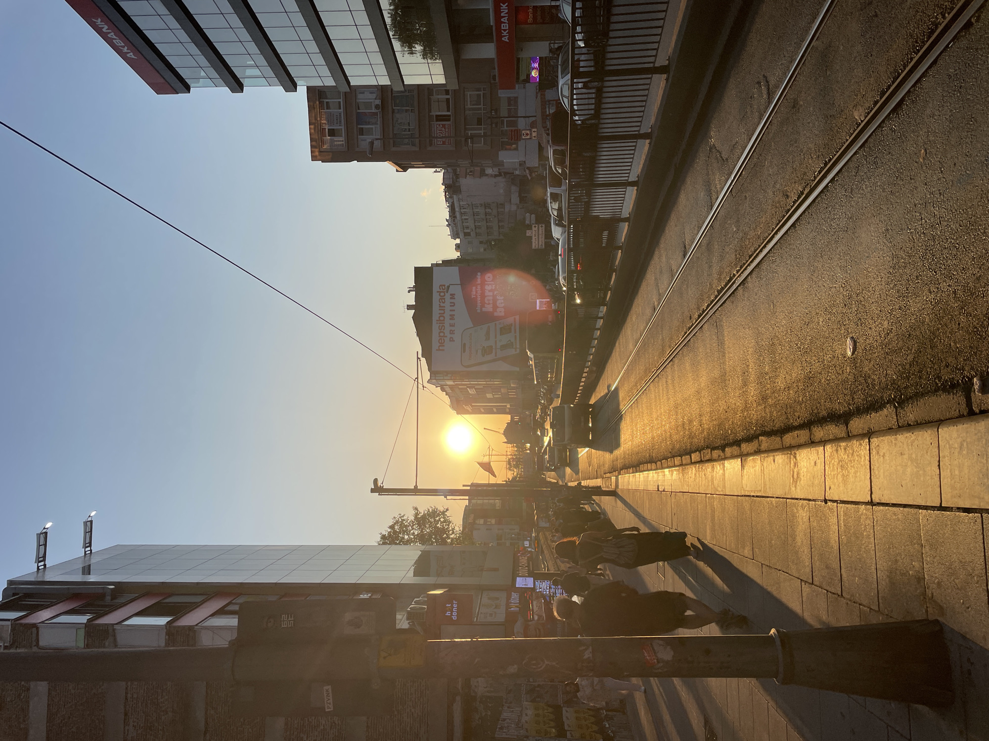 The image depicts a street scene in Kadıköy, Turkey, during sunset. The sun is low in the sky, casting a warm, golden light across the scene. The street is lined with buildings, some modern with glass facades, and others older brick structures. Tram tracks run down the center of the street, and a tram is visible in the distance. Several cars are also present, suggesting traffic. Pedestrians are walking on the sidewalk, silhouetted against the bright sunlight. The overall atmosphere is one of a busy urban environment bathed in the soft glow of a setting sun.