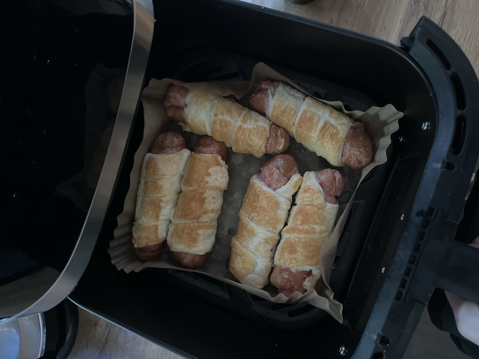 The image shows an air fryer with six cooked pigs in a blanket arranged on parchment paper. The air fryer is black and has a handle on the right side. The pigs in a blanket have a golden-brown crust. The countertop appears to be made of wood. The location of the scene is not discernible from the provided image.
