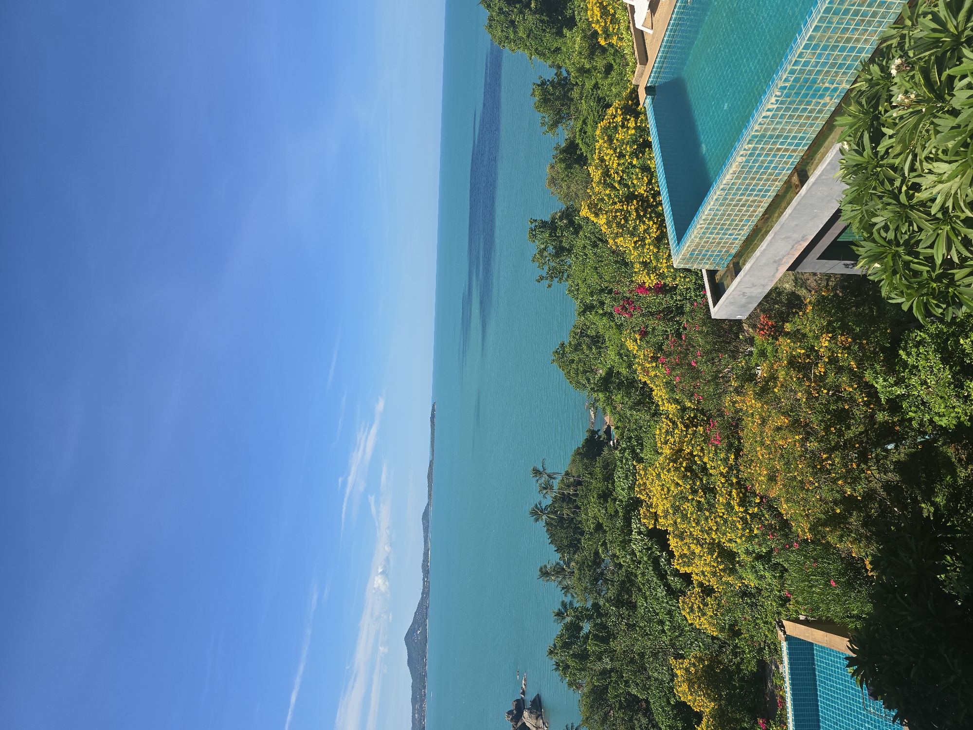 An elevated, wide shot captures a serene, tropical landscape under a bright blue sky with scattered white clouds. In the foreground, two infinity pools with turquoise water are visible, one partially in the lower left and another larger one dominating the right side of the frame. The larger pool features a tiled edge and a surrounding concrete structure. Lush, green foliage with vibrant yellow and pink flowering bushes covers the hillside, creating a dense, natural border. Beyond the foliage, a vast expanse of calm, turquoise ocean stretches to the horizon. In the distance, a chain of green, mountainous islands is silhouetted against the sky. The overall scene evokes a sense of luxurious tranquility in a remote, tropical setting.