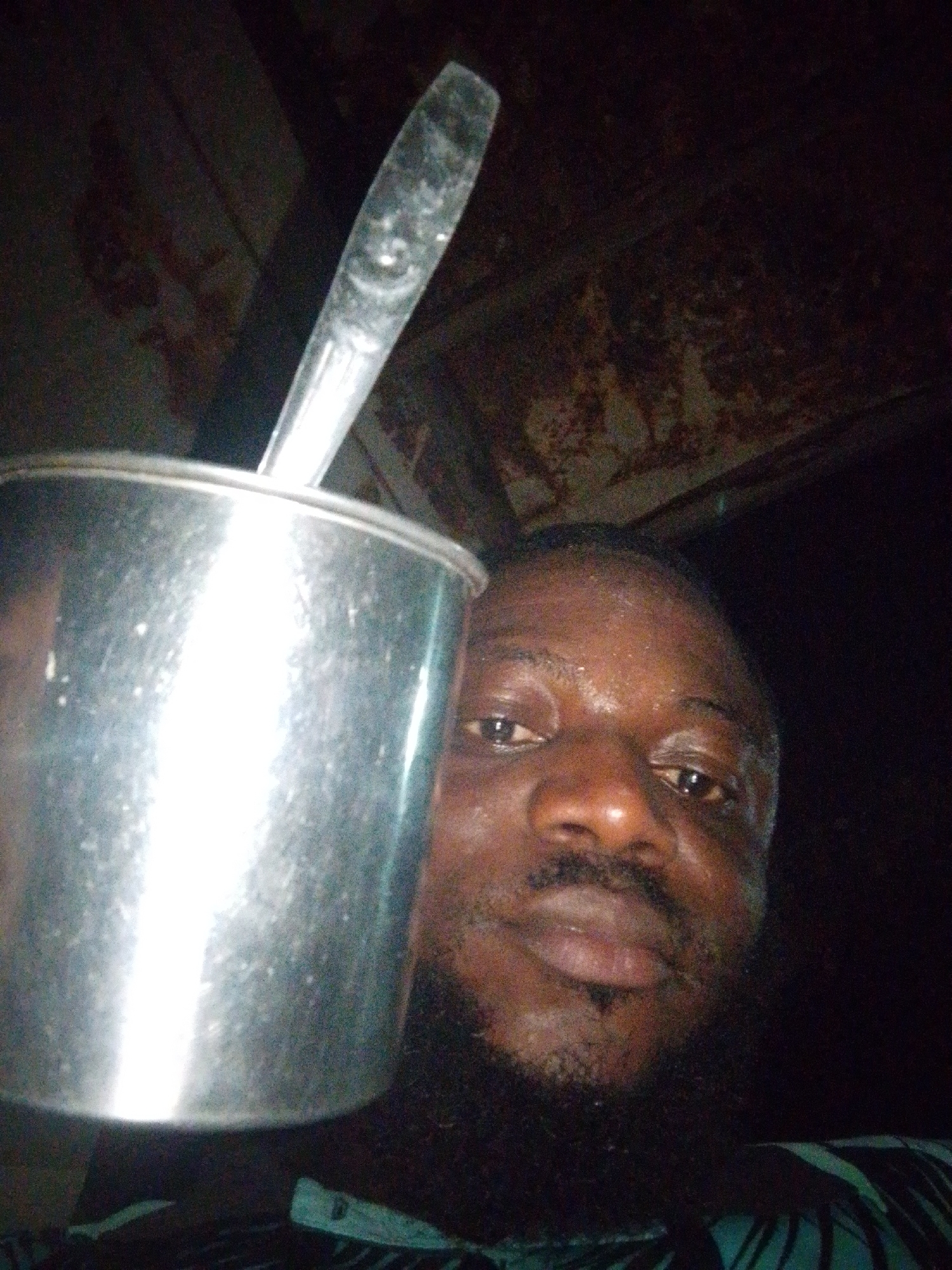 A man with dark skin, a beard, and a mustache holds a metal cup with a spoon in it. The cup reflects light. The background is dark and indistinct, but appears to be a ceiling with some patterns or stains. The picture was taken indoors. I cannot determine the exact location in Lokoja, Nigeria.