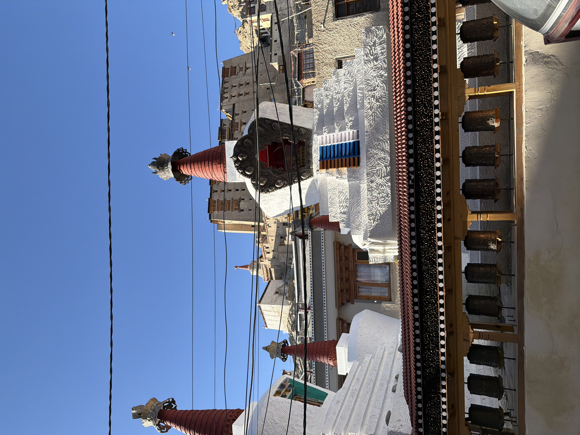 The image depicts a view of Buddhist stupas and a monastery complex, likely in Leh, India, given the architectural style and mountainous backdrop. In the foreground, two white stupas with red conical tops and decorative crowns are visible. A wooden structure with intricately carved beams and a red tiled roof runs across the bottom of the frame, holding a row of prayer wheels. Beyond the stupas and prayer wheels, the monastery buildings, constructed from stone and earth, ascend a rocky hillside under a clear blue sky. The architecture is characteristic of the Ladakh region, with multi-story structures and narrow windows. Power lines crisscross the upper portion of the image, suggesting a modern presence within the historical setting. No people are visible in the frame.