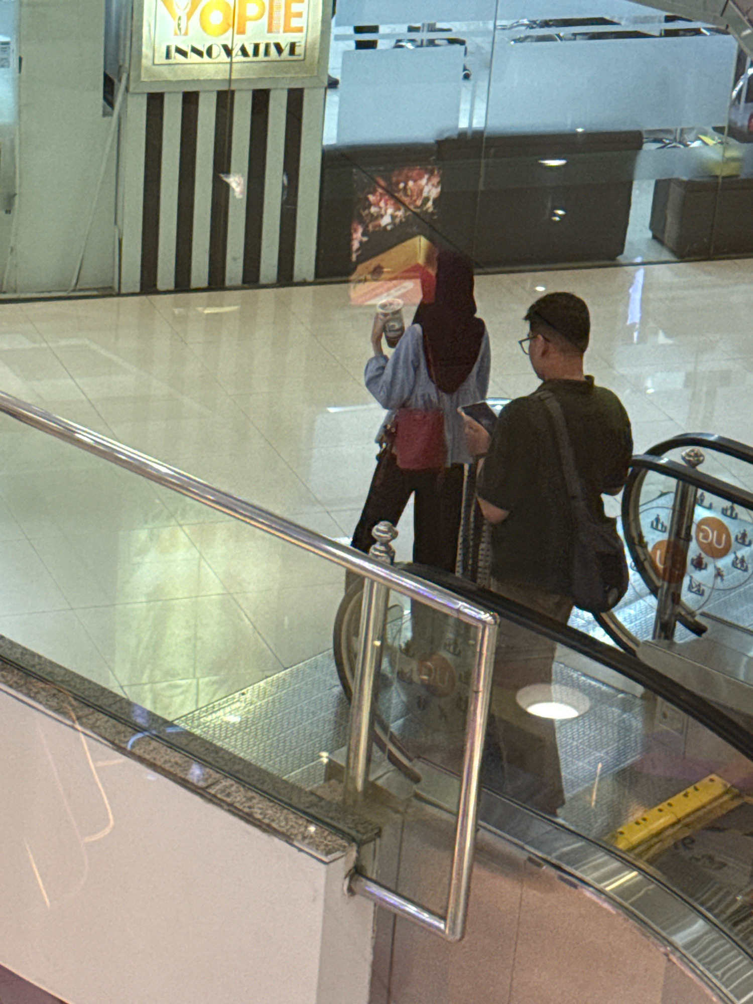 The image captures an indoor escalator scene, likely within a mall in Surabaya, Indonesia, given the context provided. Two individuals are prominently featured. On the left, a person wearing a dark hijab and a light blue shirt is holding a beverage cup in their left hand and looking down. They are also carrying a small red bag on their right side. To the right of this individual, a person wearing a dark cap, glasses, and a dark green t-shirt is looking at a mobile device they are holding. A dark bag is slung across their right shoulder. Both individuals are positioned on the escalator. The background shows a modern interior with reflective surfaces, a striped pillar with a "YOPIE INNOVATIVE" sign, and further escalators. The overall lighting suggests a well-lit commercial space.