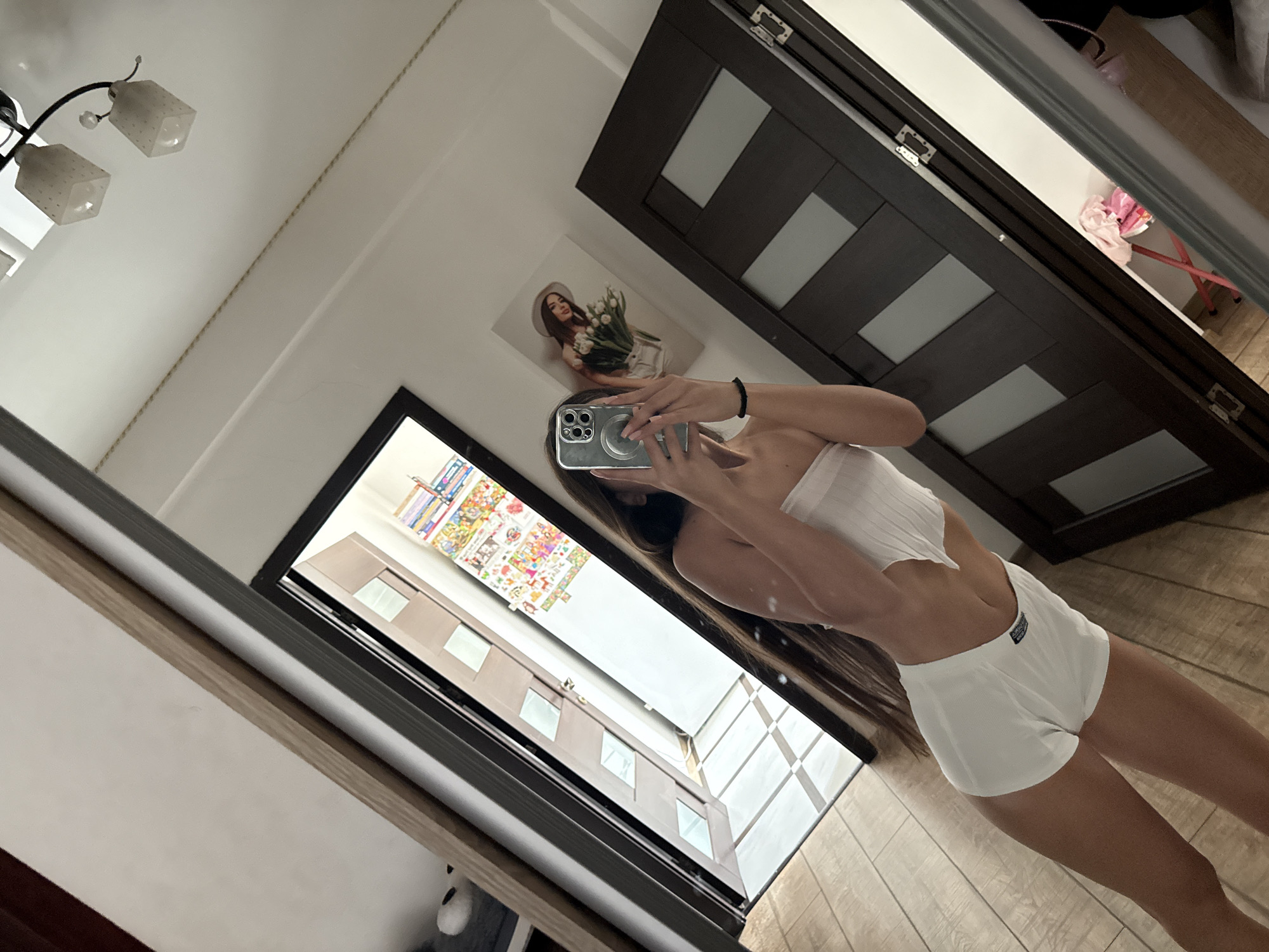 The image captures a young woman with long brown hair standing in what appears to be a well-lit room, possibly a bedroom or dressing area. She is wearing white shorts and a white bandeau top, holding a smartphone in her right hand, which is angled to take a selfie in a large mirror. The mirror reflects the room, showing a dark wooden door with frosted glass panels, a framed picture of a woman holding tulips on the wall, and a ceiling light fixture. The floor is made of light-colored wooden planks. To the left of the woman, a doorway reveals a brightly lit hallway with patterned tiles on the floor and several shelves stacked with colorful items, possibly books or toys. The overall impression is a personal, indoor space, likely a residential apartment. The specific location in Chișinău, Moldova, cannot be determined from the visual information provided in the image.