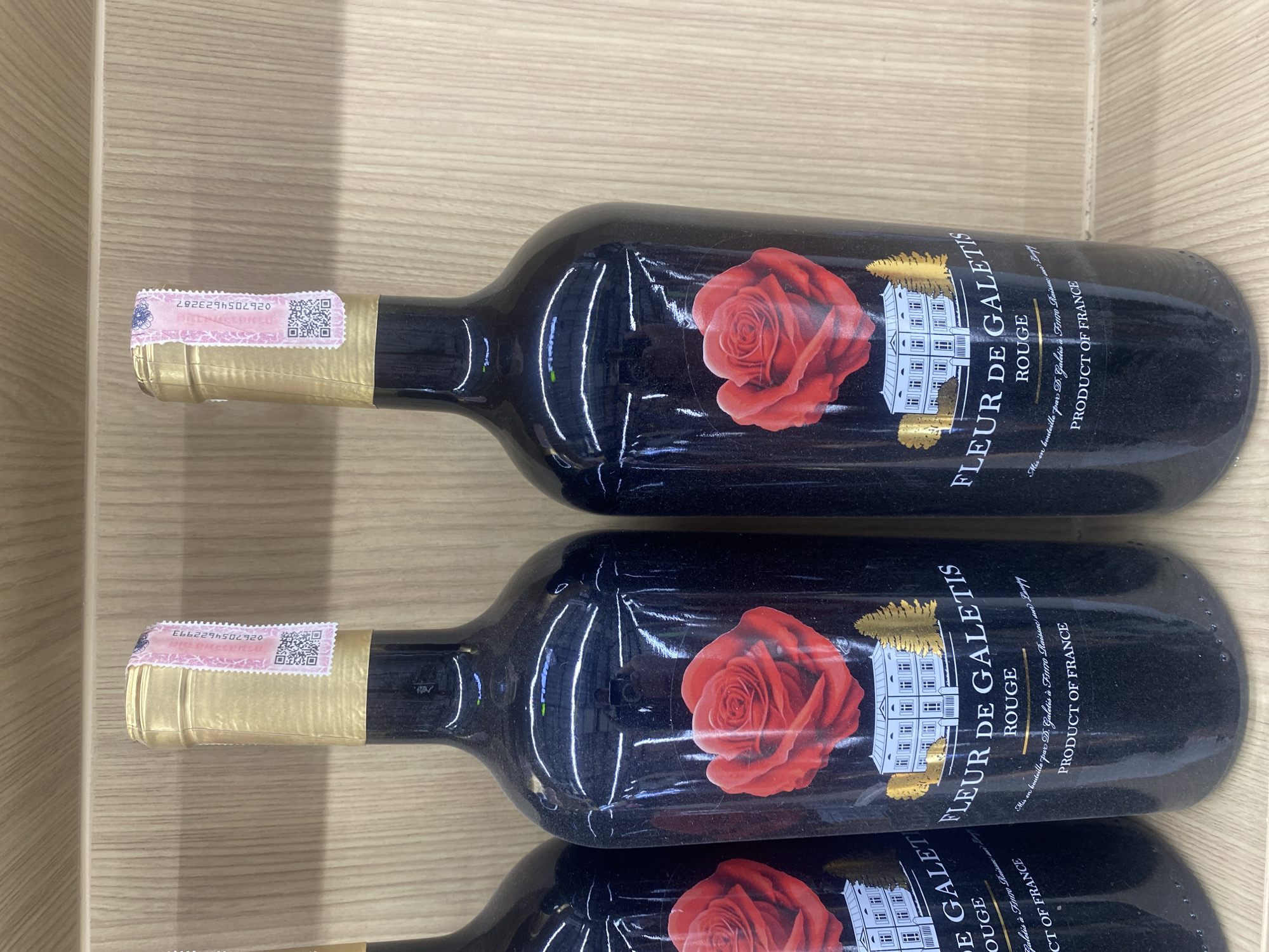 Two dark-colored wine bottles are prominently displayed, lying horizontally on a light wooden surface, possibly a shelf or crate. A third bottle is partially visible at the bottom edge of the frame. Each bottle is made of dark, almost black, glass and features a gold foil capsule covering the cork and neck.
The main label on each bottle is black with a striking red rose illustration on the left side. To the right of the rose, a white, château-like building with multiple windows is depicted. The primary text on the label reads "FLEUR DE GALETIS" in a stylized font, followed by "ROUGE". Below this, smaller text states "Mis en bouteille par D. Galetis à F11170 Raissac sur Lampy" and "PRODUCT OF FRANCE", indicating the wine's origin.
Around the neck of each bottle, a pink and white security or tax stamp is affixed over the gold foil. These stamps feature numerical codes and QR codes. The visible numerical codes are "0267054622993" on the top bottle and "0267054623287" on the middle bottle. The bottles appear to be new and some have minor reflections or dust on their surfaces. The setting is indoors, with the wooden background suggesting a retail display or storage area in Moo 1, Thailand. No human activity is depicted.