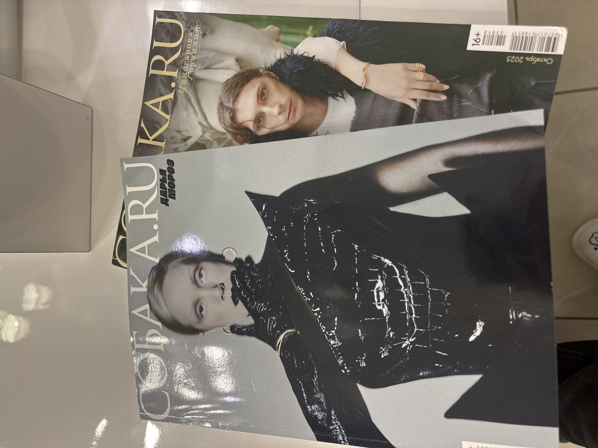 The image shows two copies of the Russian magazine "СОБАКА.RU" (SOBAKA.RU), one partially obscuring the other. The magazine on top is from October 2025. The magazine below has "Дарья Мороз" (Darya Moroz) printed on the cover. The magazines appear to be lying on a flat surface. The environment seems to be indoors, possibly a store or a similar retail space. A portion of a shoe is visible in the background. There is no information provided to determine the exact location of the city Vsevolozhsk, Russia in the image.