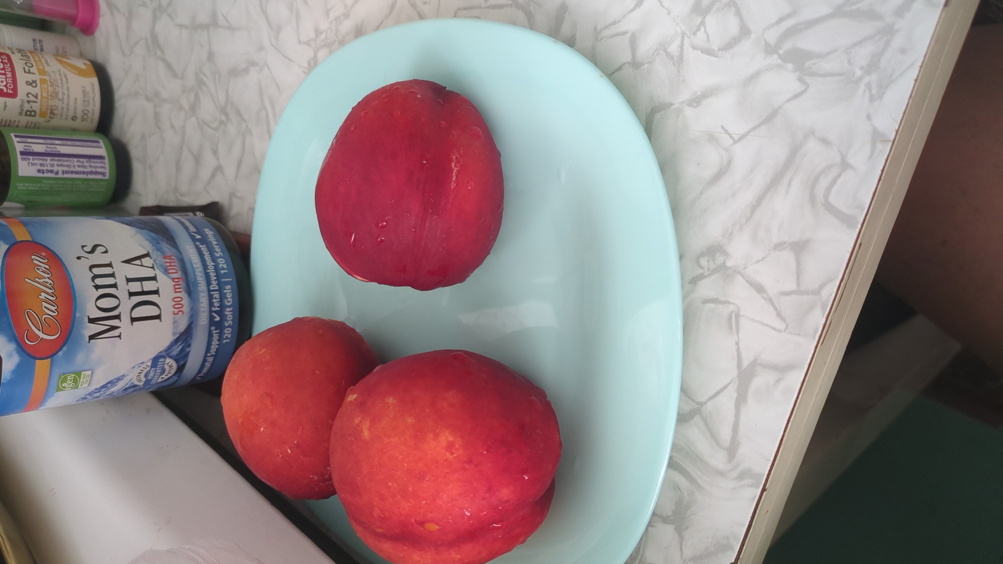 On a light blue plate, three nectarines are arranged. Two nectarines are positioned on the left side of the plate, while a single nectarine is on the right. In the background, a bottle labeled "Carlson Mom's DHA 500 mg DHA" stands upright. The plate rests on a countertop with a marbled pattern. The image was captured indoors.