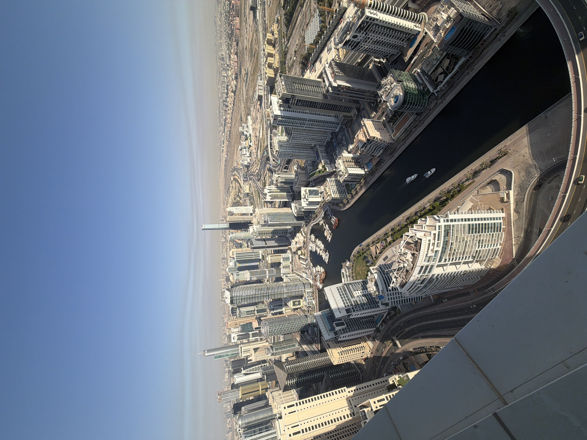 This is a high-angle panoramic view of a modern metropolis, likely Dubai, characterized by its dense collection of high-rise buildings and skyscrapers. The image is taken from a significant elevation, looking down and across the expansive urban landscape under a bright, clear sky that shows a distinct haze layer towards the distant horizon.
A dark, winding canal or waterway is a prominent feature, cutting through the city. Two white boats are visible on its surface, indicating activity. Surrounding the canal are numerous contemporary buildings with varied architectural styles, including those with extensive glass facades and others with more solid, structured exteriors. An intricate network of roads, highways, and bridges is interwoven among the buildings, with several vehicles seen traversing them. In the far left distance, a very tall, slender skyscraper stands out against the hazy sky. The bottom right corner of the frame shows part of the building or structure from which the photograph was taken, suggesting an observation deck or a high-floor window.
The scene depicts a typical daytime setting with bright sunlight, casting shadows on some buildings. There are no discernible people or legible text visible in the photograph. The overall impression is one of immense urban development and modern infrastructure.