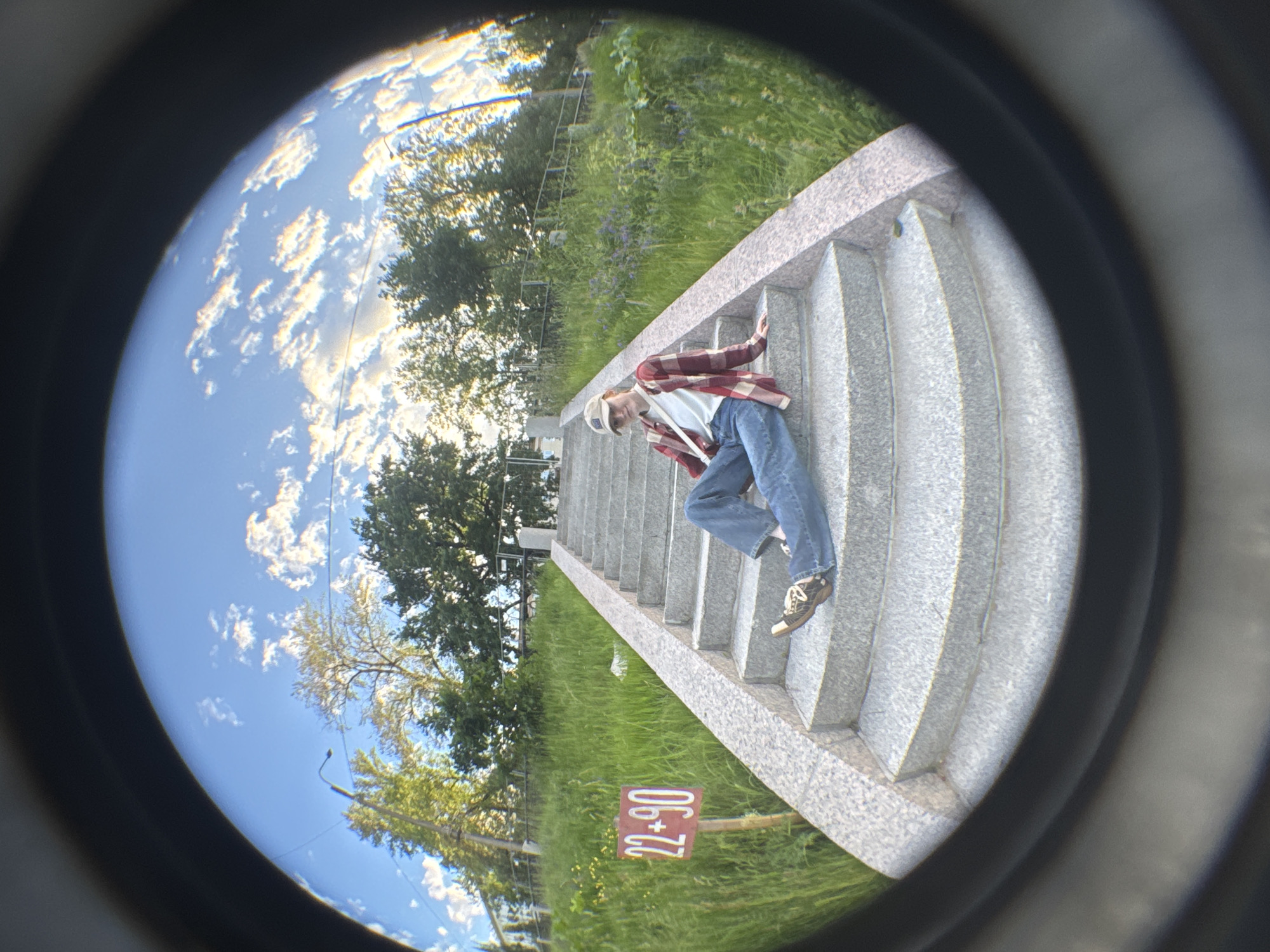 A fisheye lens captures a low-angle shot of a person sitting on an outdoor stone staircase. The person, wearing a red and white plaid shirt, blue jeans, a white t-shirt, and a beige baseball cap, is sitting facing the viewer with their legs extended. A red sign with "22+90" in white lettering is visible on the grassy embankment to the left of the stairs. The staircase is flanked by lush green grass and trees. The sky above is a clear blue with scattered white clouds, some of which are illuminated by the sun, giving them a golden hue. The overall scene appears to be set in a park or public outdoor space in Saint Petersburg, Russia, possibly near the Summer Garden given the architectural style of the stairs and the surrounding vegetation.