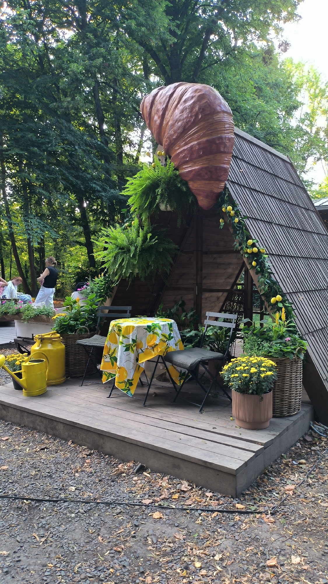 The image depicts an outdoor seating area with a wooden deck and a rustic, A-frame wooden structure in a garden setting. A large, decorative croissant sculpture is prominently displayed above the A-frame. A round table with a yellow tablecloth featuring a lemon print is set with two chairs, suggesting a cafe or a dining space. Various potted plants, including ferns and yellow chrysanthemums, are arranged around the deck. Two yellow watering cans are also present on the deck. In the background, two individuals are visible, one standing and one sitting, near a low planter box. The environment is characterized by lush green trees, suggesting a park or a wooded area. The overall scene is peaceful and evokes a whimsical, outdoor cafe atmosphere. There is no indication within the image that this location is in Lviv, Ukraine.
