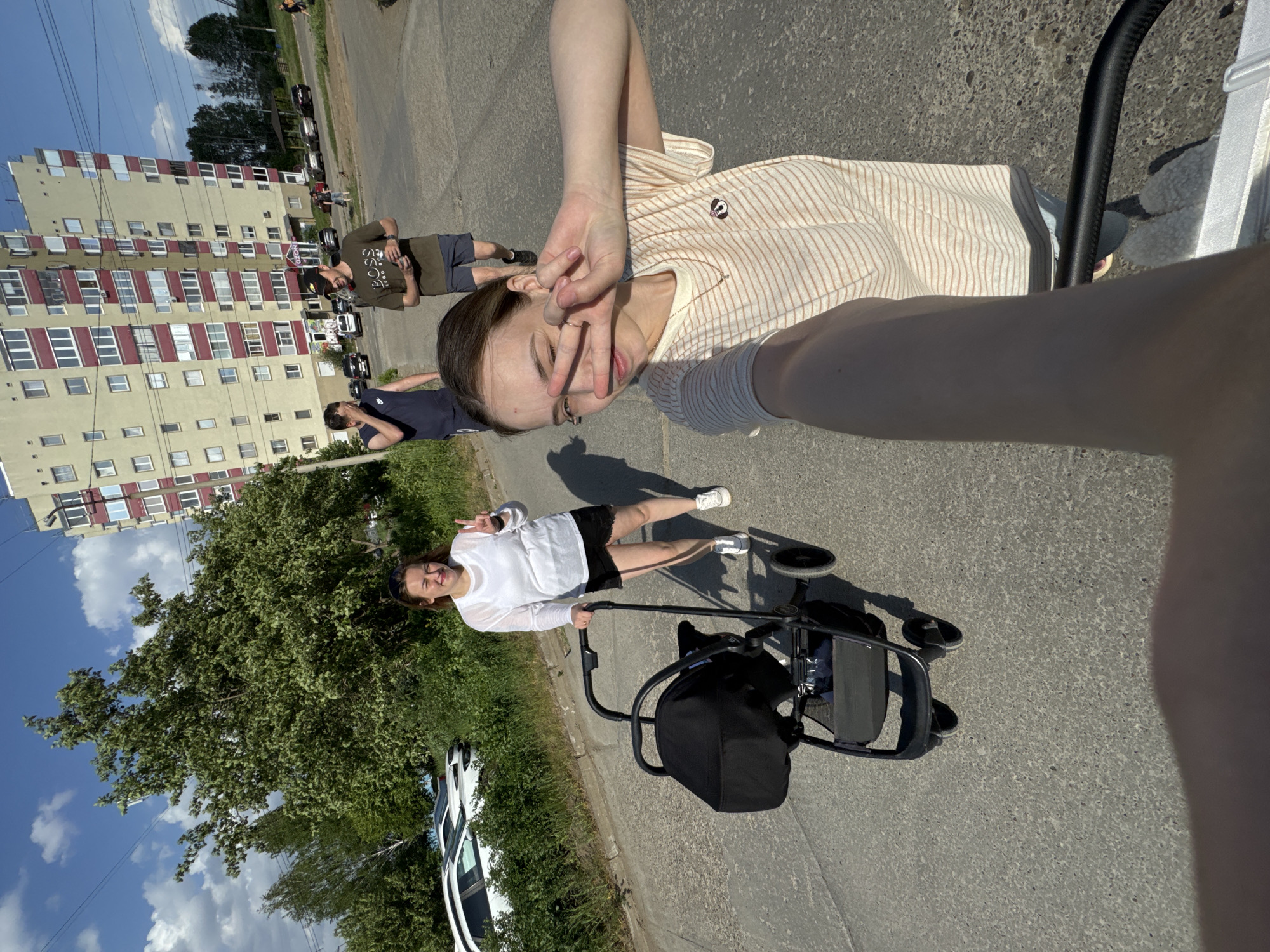 The image shows an outdoor scene in what appears to be the city of Chaikovsky, Russia, judging by the architecture. The picture is a selfie, with the photographer partially visible. The photographer is wearing a striped shirt and gesturing with their hand near their face.
In the background, another individual is seen pushing a stroller. They are wearing a white top and dark shorts and also gesturing with their hand. A white car is parked nearby, close to trees and greenery. Beyond them are more people standing near a multi-story apartment building with light-colored walls and red accents. The ground is asphalt. The sky is blue with scattered clouds.