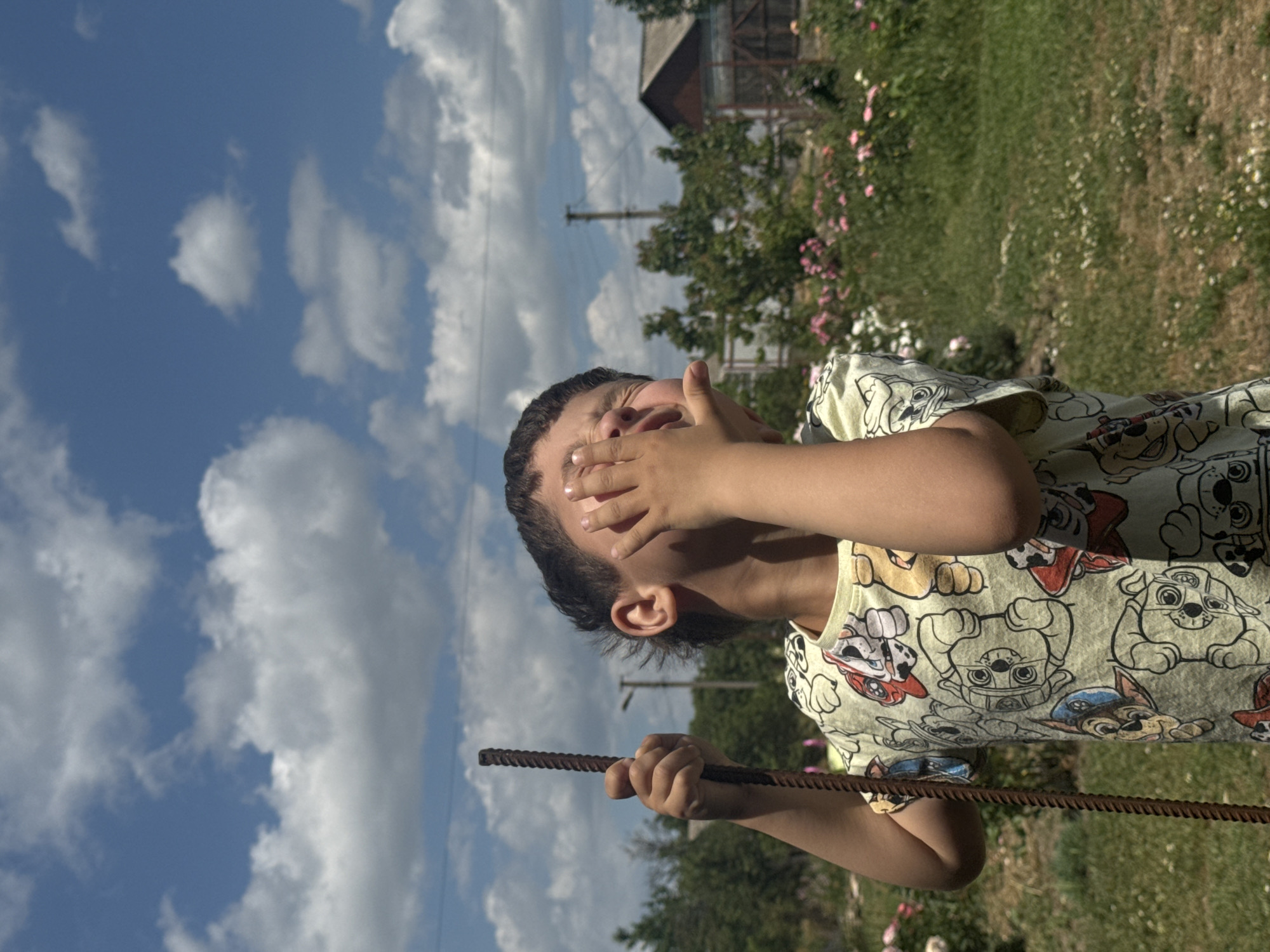 A young child with dark hair is captured outdoors in a medium shot, holding a rusty metal rod in their left hand and covering their face with their right hand. The child is wearing a light yellow t-shirt adorned with a repeating pattern of cartoon dogs. The background features a lush green lawn dotted with small white flowers and blooming rose bushes. Beyond the garden, trees and a rustic wooden house with a fence are visible under a bright blue sky filled with fluffy white clouds. The scene suggests a rural or suburban setting, possibly a backyard or garden.