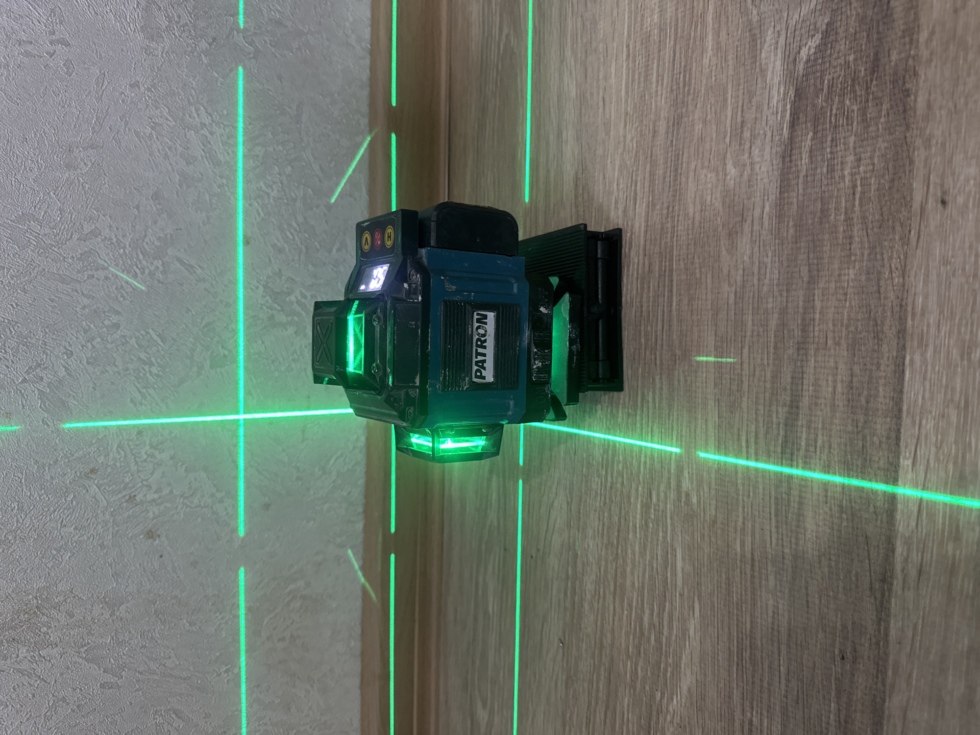 A green laser level projects intersecting horizontal and vertical lines onto two walls meeting at a corner. One wall has a textured, light-colored finish, possibly plaster or stucco, while the other wall features a wood-grain laminate. The laser level itself, a multi-faceted device with a black and teal casing, is mounted to the wood-grain wall. Visible text on the device reads "PATRON". The scene appears to be an indoor setting, likely a room or hallway, with no indication of the time of day or weather conditions. The activity is the projection of laser lines, suggesting a task related to construction, renovation, or measurement is in progress.
