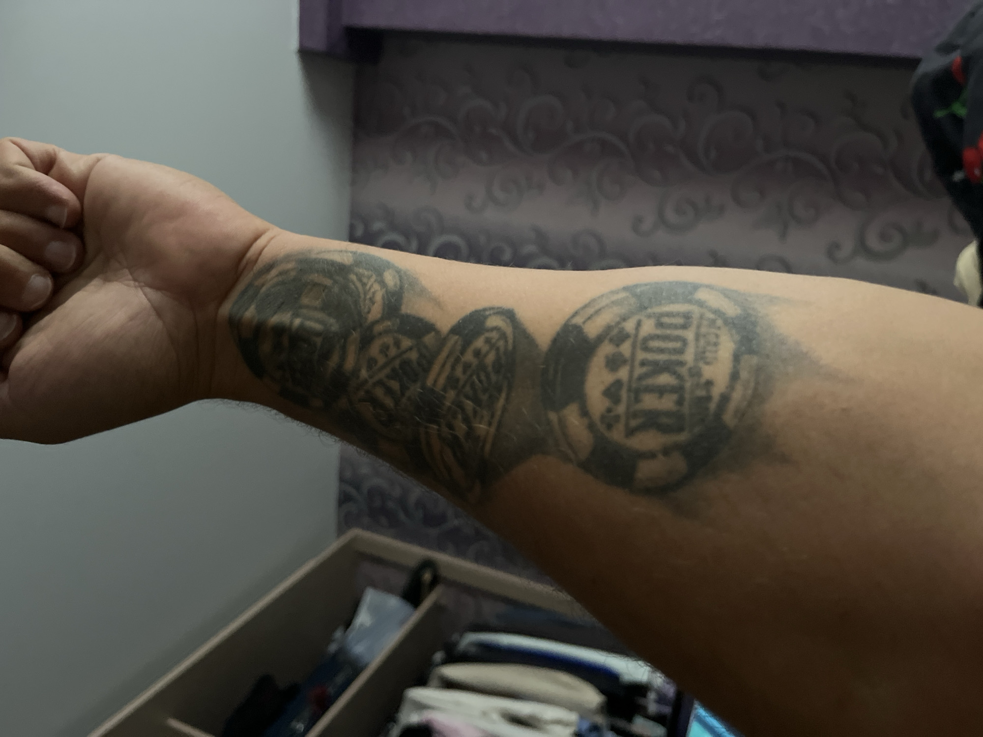 The image shows a person's forearm with several tattoos of poker chips and dice. The tattoos are black and grey and appear to be old. The person's arm is resting on a surface that is out of focus. In the background, there is a closet with clothes hanging inside and a patterned wallpaper. The lighting in the image is dim, suggesting it was taken indoors. There is no information in the image to suggest the location is the city of Minsk, Belarus.