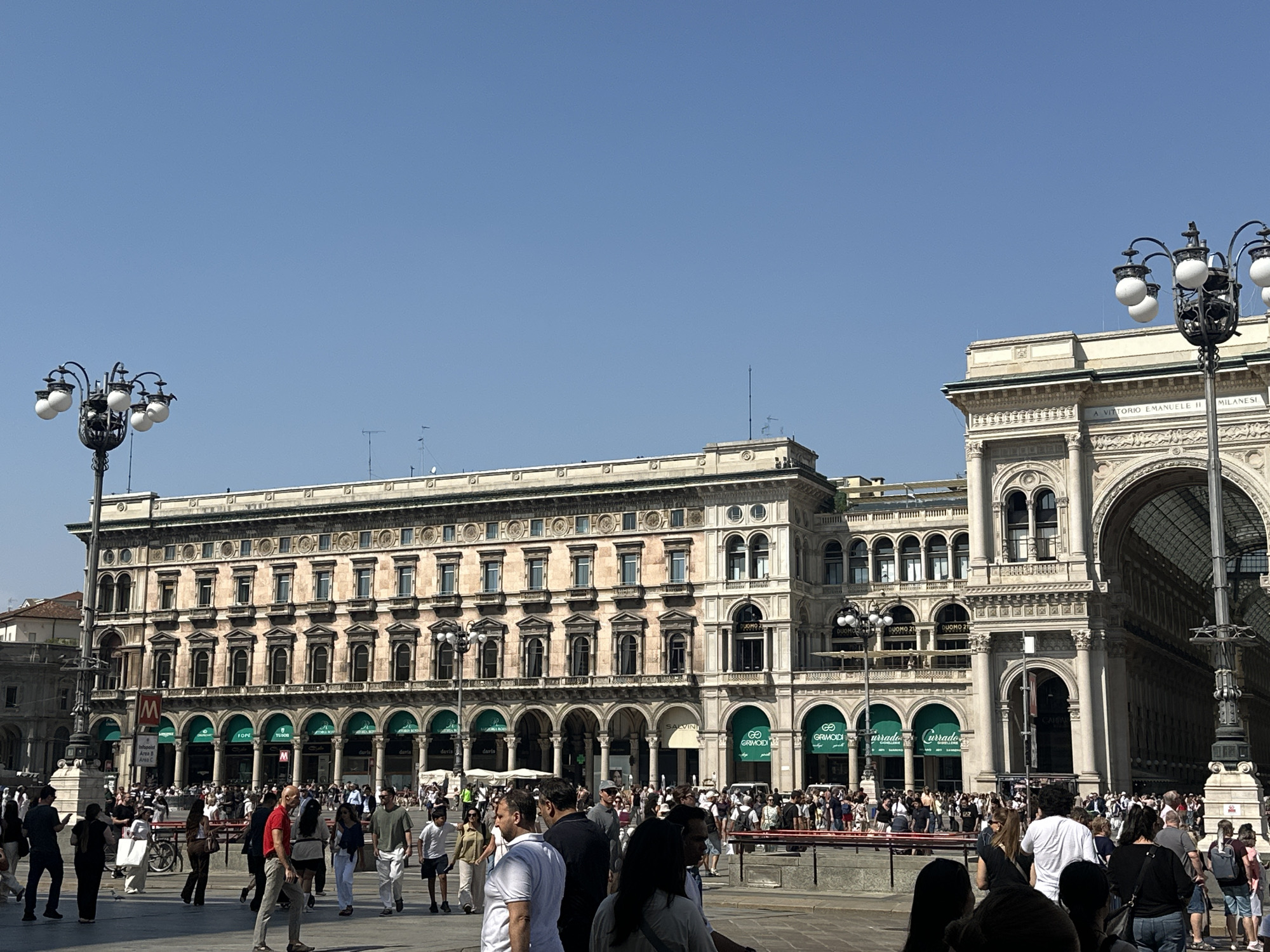 The image depicts a wide, sunlit plaza in front of an ornate, multi-story building with arched entrances and windows. A large crowd of people is milling about in the plaza, many walking in different directions. Several streetlights with multiple globes are visible, as well as trees and signage. The architecture suggests a European city, possibly a historic district. The sky is clear and blue.