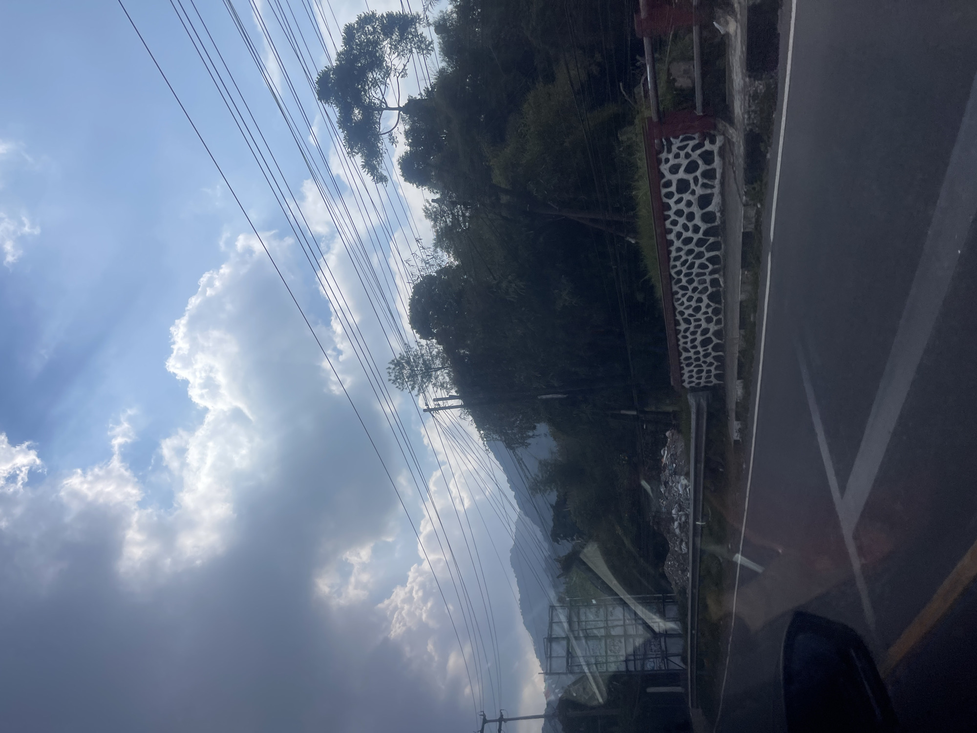This is a photo taken from inside a vehicle, looking out at a road and the surrounding landscape. The road appears to be in good condition, with a white line marking the center and yellow lines on the side. To the right of the road, there is a decorative wall with a black and white patterned design, topped with a red railing. Beyond the wall, there is a slope covered with vegetation. In the distance, there are mountains, partly obscured by trees and electrical wires that crisscross the sky. The sky is a mix of blue and dark clouds, with rays of sunlight breaking through. On the left side of the road, there is a large billboard-like structure and a pile of debris. The overall impression is a scenic view of a mountainous area, likely in a rural or semi-urban setting. The location is identified as being in the city of Ciloto, Indonesia.