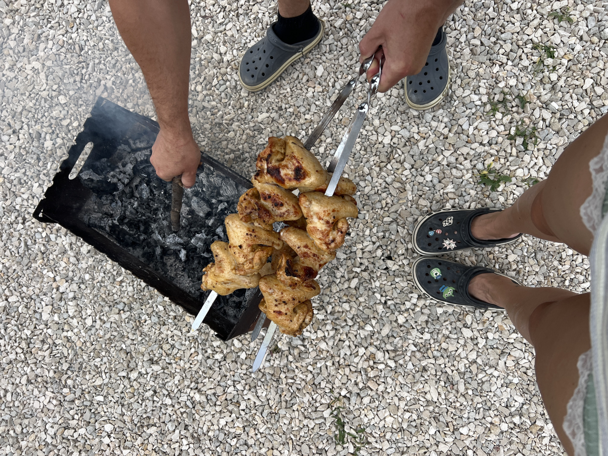 A high-angle full shot shows a kebab of chicken wings being grilled over hot coals by two individuals in an outdoor setting with a gravel ground. The chicken wings are golden brown and seasoned with black pepper. Smoke rises from the grill. One person's arm is visible holding a stick to tend the coals, while the other person's hands are shown holding tongs to adjust the chicken. Both individuals are wearing dark-colored Crocs. The ground is covered with small, light-colored gravel.