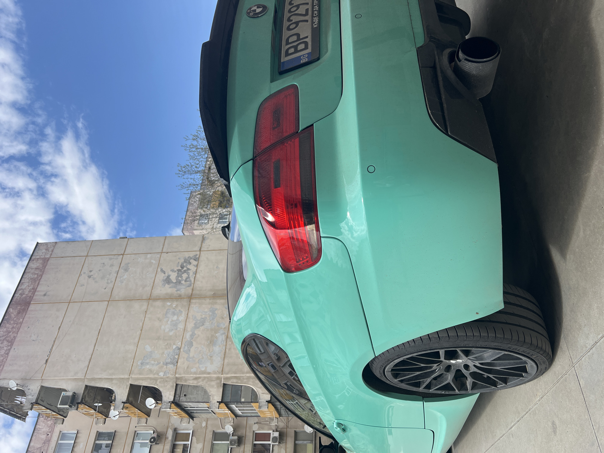The image, presented in a vertical orientation, captures the rear three-quarter view of a striking teal (mint green) BMW coupe, likely an M-series, parked in an urban environment. The car's vibrant color stands out against the more subdued background.
The BMW's rear features a prominent red LED taillight, a black lip spoiler on the trunk, and an aggressive black rear diffuser. A large, single black exhaust tip is visible on the right side. The circular BMW logo is present above the license plate. The Bulgarian license plate, "BG BP 9297", is clearly visible, with additional text "КЪДЕ СИ ДА ПРИ..." (translating to "WHERE ARE YOU TO COME...") partially legible. The car is fitted with multi-spoke black aftermarket wheels, and the tire brand "SOLMAX" is discernible on the sidewall.
The background consists of a tall, weathered concrete apartment building with a panelized facade, typical of Eastern European architectural styles. The building's surface shows signs of age and wear. It has numerous windows, many with small balconies, some equipped with awnings, satellite dishes, and air conditioning units.
The scene is set outdoors during a bright, sunny day with a clear blue sky adorned with scattered white clouds. The shot is taken from a low angle, looking upwards at the car's rear and the building. The car appears to be stationary, likely parked on a street or a paved area in Plovdiv, Bulgaria.