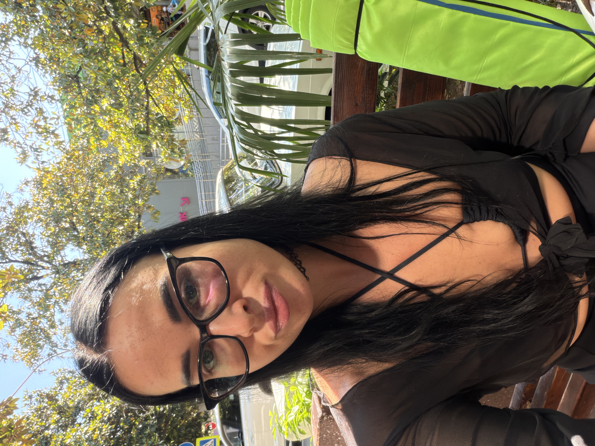 A woman with dark hair and glasses takes a selfie while sitting on a wooden bench outdoors. She is wearing a black sheer top over a black bikini top. Behind her, trees with yellowing leaves and a building with a pink sign are visible. A bright green rolled-up object is next to her on the bench. The scene appears to be a public area in Sochi, Russia, with cars and pedestrian walkways in the background.