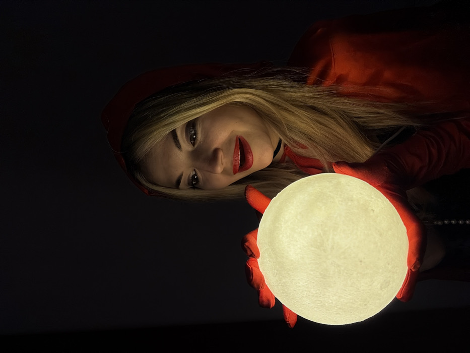 A fair-skinned woman with blonde hair, dressed in red, poses with a glowing orb. She wears a red hooded cloak and matching gloves, her face illuminated by the orb she holds. The background is dark, emphasizing the light emanating from the sphere. Her makeup includes red lipstick, complementing her outfit. A thin black choker adorns her neck, and a string of pearls is visible.