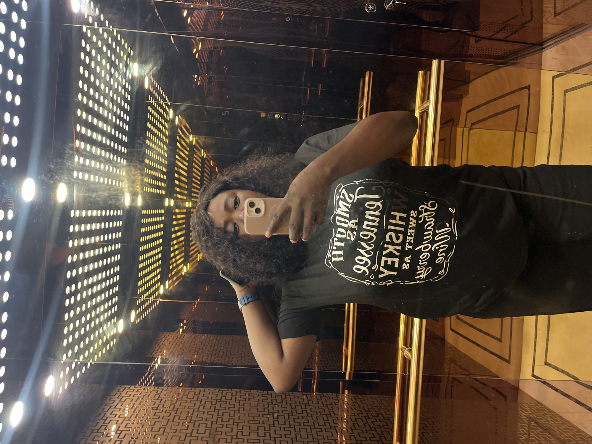 A Black woman is taking a selfie in an elevator. She is wearing a black t-shirt with white text that reads "Smoother Than Bourbon, Sweeter Than Whiskey," and black pants. She has curly black hair and is wearing a blue watch on her left wrist. The elevator has mirrored walls, a golden railing, and a patterned ceiling with lights. The floor has a geometric pattern. The elevator appears to be luxurious, possibly in a hotel or a high-end residential building in Abuja, Nigeria.