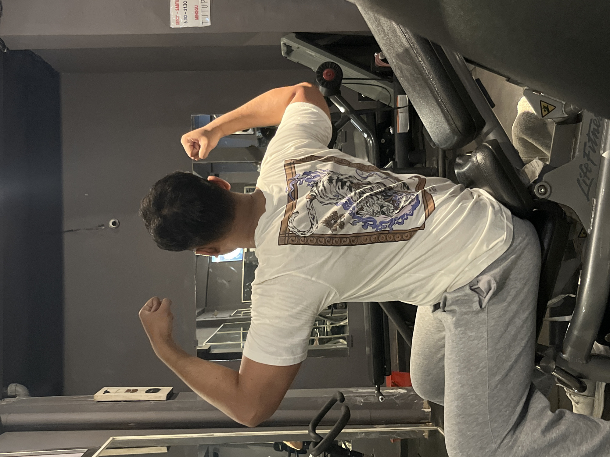 A man is seated on a weight machine in a gym. He is wearing a white t-shirt with a graphic design on the back and grey sweatpants. The man is flexing his biceps, with his arms bent and fists clenched. The gym is dimly lit with grey walls and a dark ceiling. There are other weight machines and mirrors visible in the background. A sign on the wall indicates gym hours, with "SENIN-SABTU 06.30-21.30" and "MINGGU TUTUP" (Monday-Saturday 06:30-21:30 and Sunday Closed), suggesting the location is in Indonesia. The "Life Fitness" logo is visible on the weight machine. The overall scene depicts a man engaged in weight training.