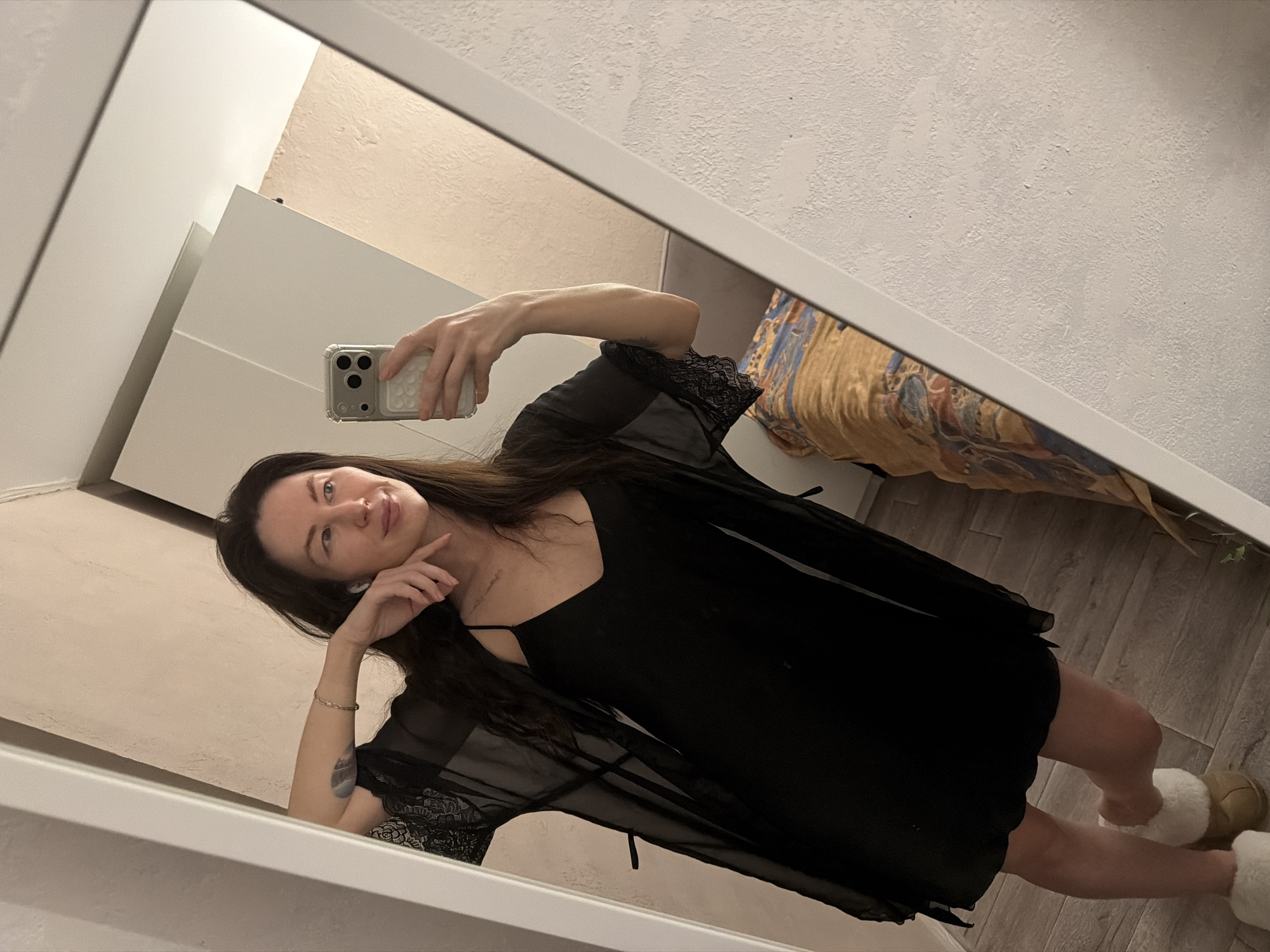 A woman is taking a selfie in a mirror. She is wearing a black dress with a sheer black robe over it, and fuzzy slippers. Her dark hair is long and loose. She is resting her head on her hand and smiling slightly. The background includes a white dresser, a wall with textured white paint, and a bed with a patterned cover. The location is likely an apartment in Perm, Russia.