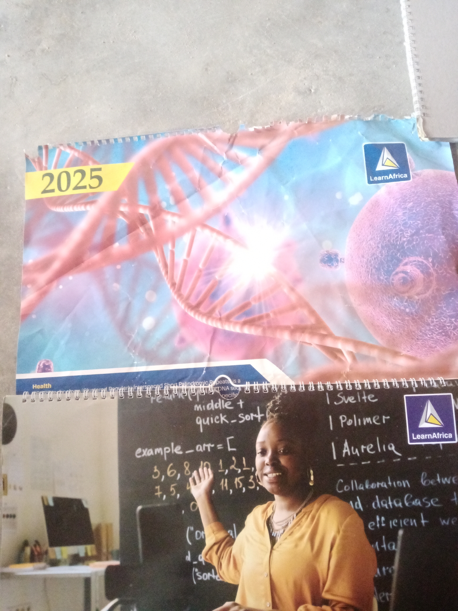 This image depicts a calendar for the year 2025, featuring a DNA helix as its cover design. Below this is a black chalkboard with handwritten text, possibly related to computer science or data. A young Black woman, wearing a yellow top and a necklace, is positioned in front of the chalkboard, with her hand raised as if explaining something. The background shows a blurred office or classroom environment with a desk, computer, and other items. The calendar also displays a "LearnAfrica" logo. While the image is visually clear, there is no discernible information or visual cues within the image that indicate its specific location in Jalingo, Nigeria.