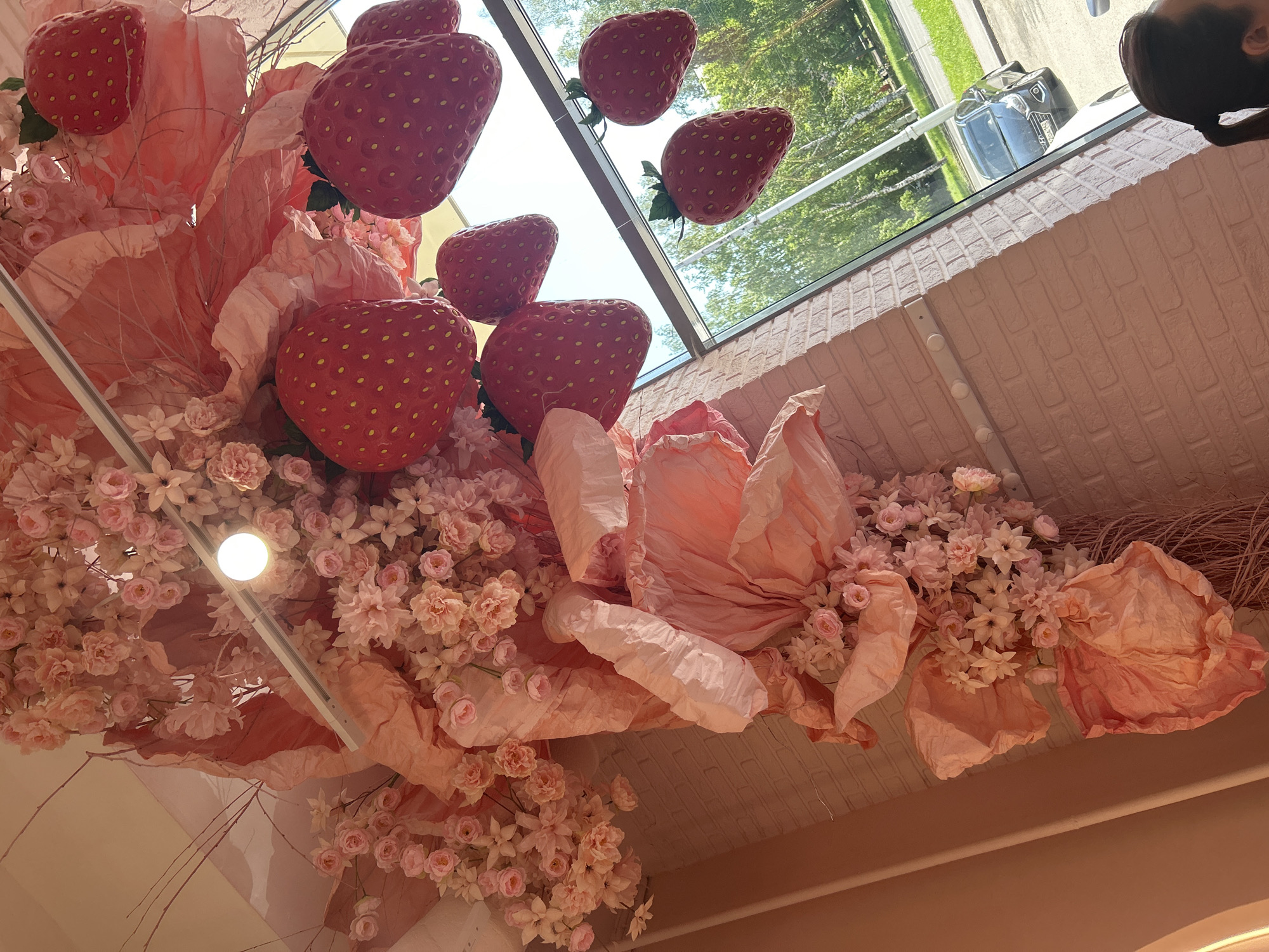 A ceiling decoration features oversized artificial strawberries interspersed with clusters of pink artificial flowers and large, crinkled pink paper elements. The strawberries are bright red with yellow seeds. The flowers are a mix of roses and other bloom shapes in various shades of pink. The paper elements are large and dramatic, adding a sense of volume to the display. In the background, through a large window, a street scene is visible with green trees, a sidewalk, and a dark-colored car parked on the side of the road. A section of a pink brick wall is visible on the left side of the image, adorned with small white lights or fixtures.