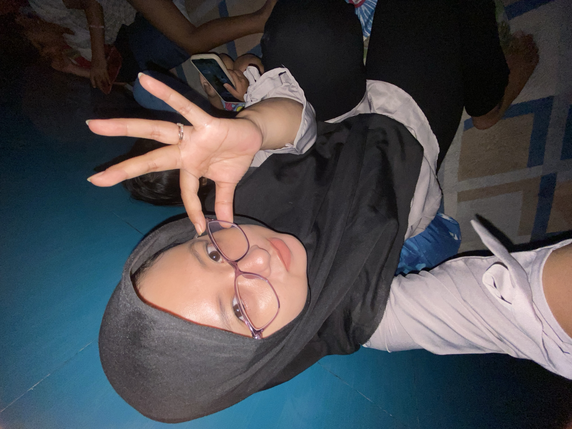 An indoor scene in Gelam, Indonesia, features a young, light-skinned woman positioned centrally, looking directly at the camera. She wears a black hijab, a light lavender long-sleeved shirt, black trousers, and light purple-rimmed eyeglasses with light pink lipstick. Her right hand is raised, palm outward with fingers spread, holding the right temple of her glasses. A silver-toned ring is visible on her right ring finger. To her immediate left is a dark teal wall. To her right and behind her, the dark hair and partial form of another person are visible. Further to the right, additional individuals are dimly seen, one holding a white smartphone, and another with bare legs and feet. All subjects are seated on a patterned mat or rug with blue, grey, and white geometric designs. The lighting is dim, suggesting an evening or night setting, with artificial light illuminating the central figure. The woman's bare left foot is visible on the mat.