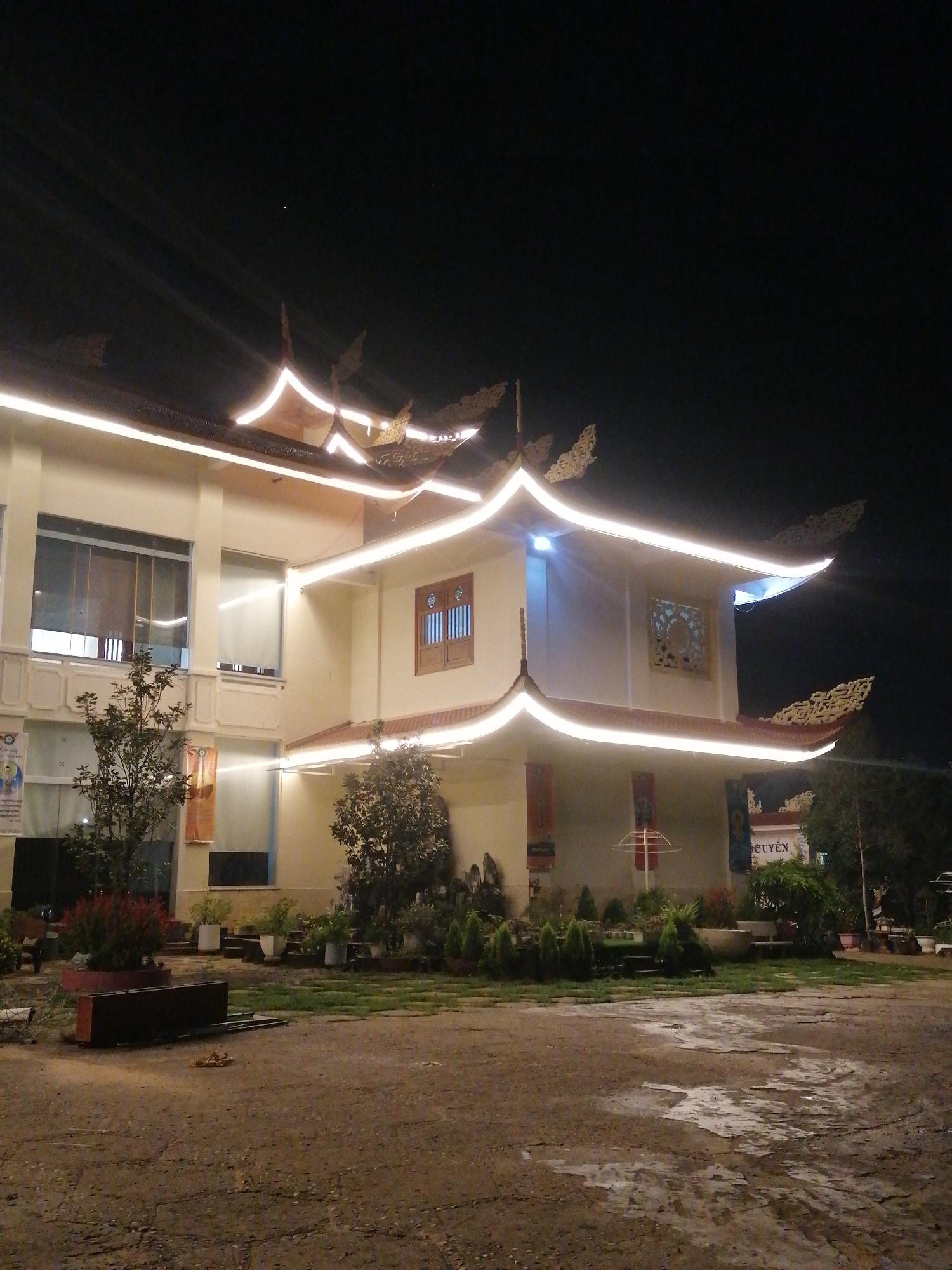 The image shows a building illuminated at night in Da Lat, Vietnam. The building has a multi-tiered roof with upturned eaves decorated with white lights. Decorative elements adorn the edges of the roof. The building is two stories in height with windows on both levels. Plants and shrubs are in front of the building. The foreground is a paved area. The sky is dark.