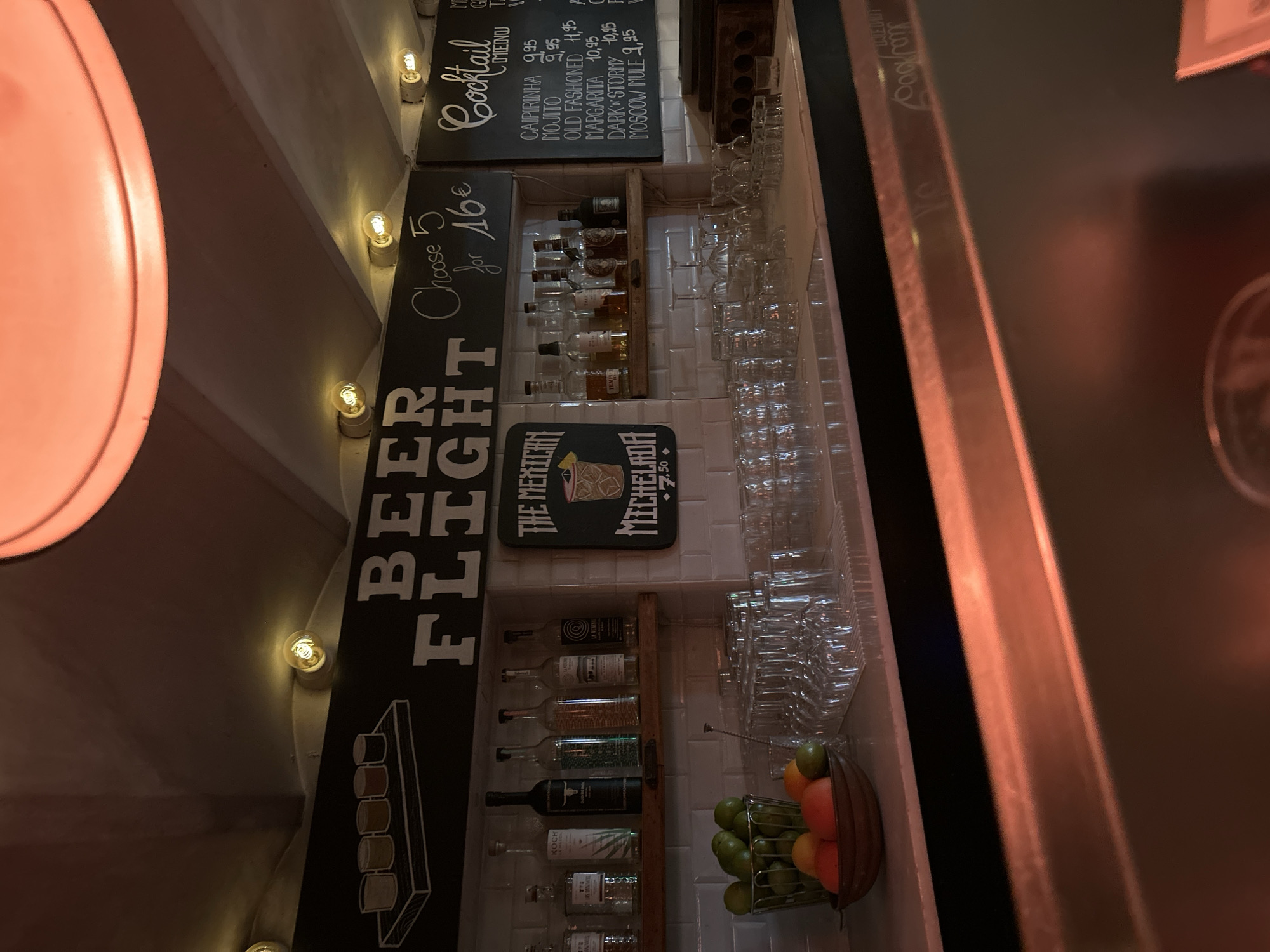 The image shows a dimly lit bar counter area, possibly a food truck or a small establishment, featuring a menu board, bottles of alcohol, glassware, and fruit. The menu board on the left displays "BEER FLIGHT" with an offer for "Choose 5 for 16€." To the right, another menu board lists "Cocktail" options, including "CAIPIRINHA 9,95," "MOJITO 9,95," "OLD FASHIONED 11,95," "MARGARITA 10,95," "DARK N' STORMY 10,95," and "MOSCOW MULE 9,95." There are shelves stocked with various bottles of spirits, and a bowl of limes and oranges sits on the counter, alongside stacks of clean glassware. The overall ambiance suggests a casual bar setting. The location is identified as Barcelona, Spain.