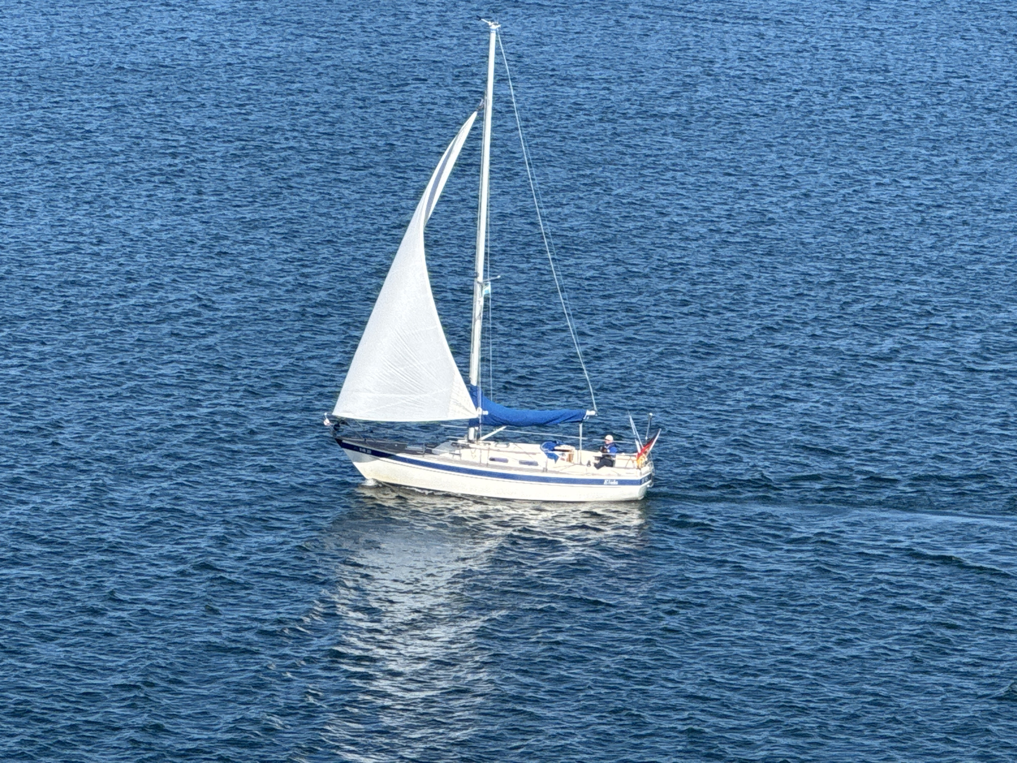 An aerial view captures a white sailboat with blue trim gliding across a deep blue, rippled body of water. The boat's main sail is fully deployed and catches the light, while a smaller blue sail is furled. A single person, wearing a light-colored hat, is visible on the deck of the sailboat, positioned near the stern. Two flags, one red and one with a yellow and blue design, are attached to the stern. The sailboat leaves a visible wake behind it, and its reflection is cast on the water's surface. The environment is entirely water, with no land features visible in the frame. This scene is consistent with the waters surrounding Karlskrona, Sweden, a coastal city known for its archipelago and naval history.