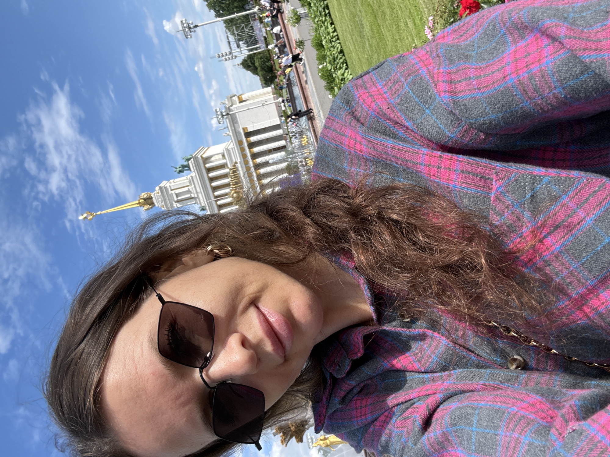 A woman is taking a selfie outdoors, with a building and a grassy area in the background. She has long, brown hair and is wearing a pink and grey plaid shirt and dark-rimmed sunglasses. A gold-colored chain is visible across her shirt. The sky is blue with white clouds, suggesting it is daytime and fair weather. The background shows a large, ornate white building with a golden spire, which is characteristic of architecture found in Moscow, Russia. The grassy area and what appear to be flower beds suggest a park or an exhibition ground. The woman's expression is relaxed and pleasant, with a slight smile. There is no visible text in the image.