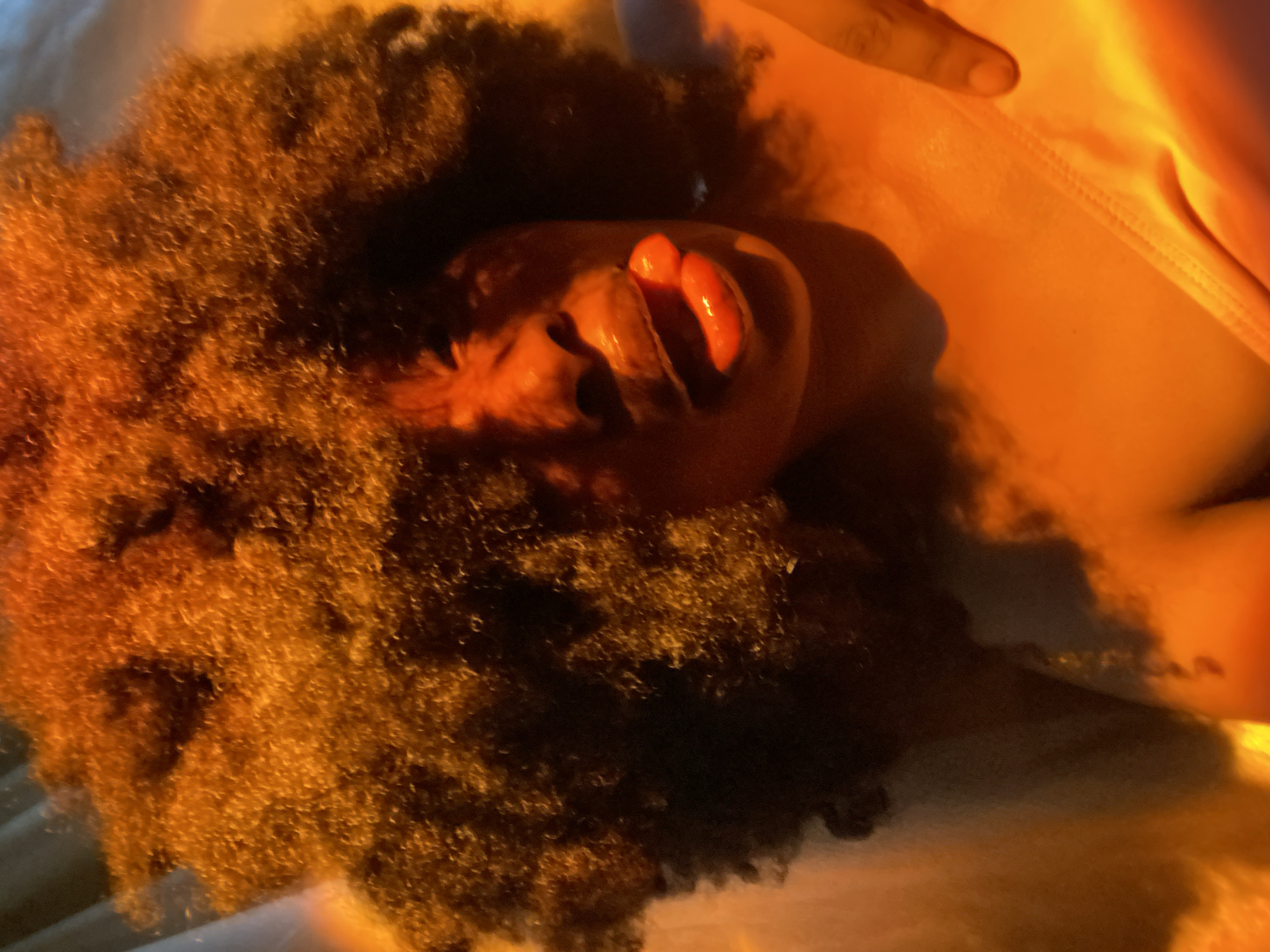 A person with afro-textured hair lies supine. The head is positioned centrally, with the face tilted upwards. A finger rests on the chest. A garment, possibly a tank top, is visible. The scene is bathed in warm, orange light, possibly from a sunset or artificial light source. The location and actions in the city of Iranla, Nigeria cannot be determined from this image.