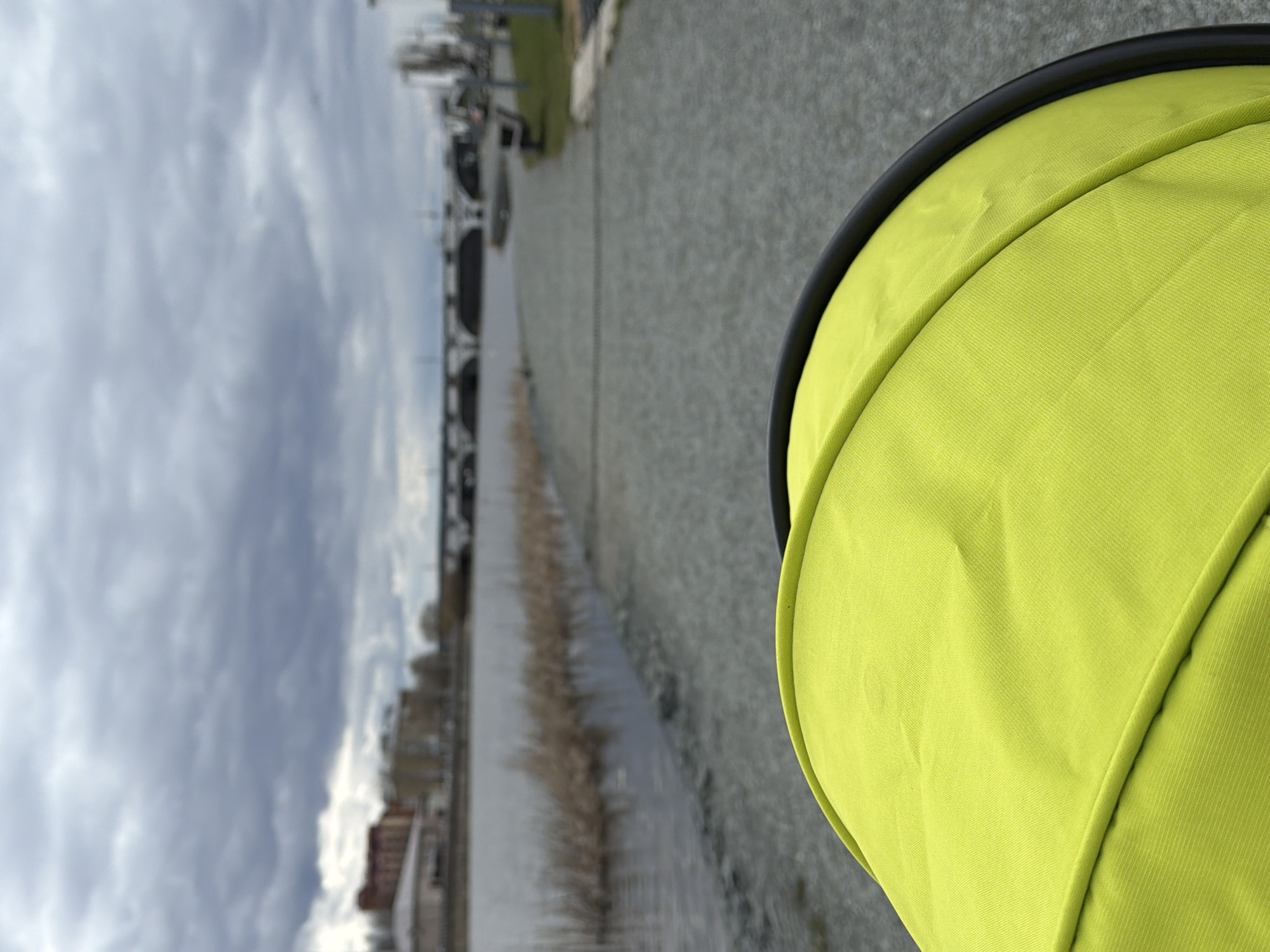 The image depicts an outdoor scene in Votkinsk, Russia, likely taken from the perspective of someone pushing a stroller or pram. A significant portion of the lower right foreground is filled by a bright lime green fabric canopy with a black rim, consistent with a stroller hood.
Stretching into the distance, a paved path or road runs parallel to a body of water, possibly a canal or river, located on the left side of the frame. The water appears calm, and some reeds or dry vegetation can be seen along its edge. In the mid-ground, a substantial structure, resembling a bridge, dam, or sluice gates, spans across the waterway. Beyond this structure, various buildings are visible in the distant background, hinting at an urban or industrial setting. The sky above is completely overcast with dense grey clouds, suggesting a cloudy daytime environment. No specific time of day is discernible, and no individuals or visible text are present in the image.