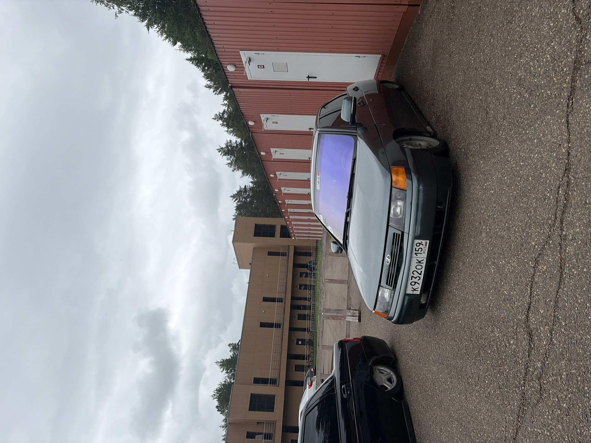 The image depicts an outdoor scene in the city of Прикамский, Russia, on an overcast day. The sky is predominantly gray with dense cloud cover.
In the foreground and mid-ground, two modified sedans are parked on a cracked asphalt surface. The car on the right is a dark green Lada 110 series, heavily lowered with extreme negative camber on its wheels, a common "stance" modification. It has a purple-blue tint strip across the top of its windshield with "БПАН" and "PUSH FAMILY" stickers visible. Its Russian license plate reads "K 932 OK 159". The car on the left, partially visible, is a black sedan, also a Lada 110 series or similar, featuring a similarly lowered suspension with its rear wheel tucked into the fender.
In the background, to the left, stands a multi-story building constructed of light brown material, possibly stucco or panels. It features numerous dark, rectangular windows and some upper-level balconies with railings. Further right, a long row of dark red corrugated metal units, resembling storage containers or temporary structures, extends across the mid-ground. Each unit has a white rectangular door with a black handle, and small white light fixtures mounted above. Door "13" is clearly visible on the unit furthest to the right.
Behind both the brown building and the red units, a dense line of coniferous trees forms the horizon. In the distance, between the brown building and the red units, a white car is parked, and several bicycles are lined up against a low fence. The ground is a dark, rough asphalt surface with visible cracks and minor debris. No identifiable individuals are performing actions in the scene.