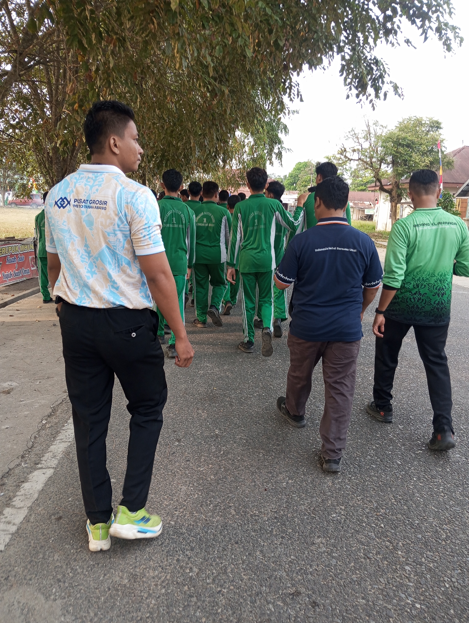 The image depicts a group of people walking on a paved road in Pematang Reba, Indonesia. The foreground features a man from the back, wearing a light blue and white patterned shirt and black pants. He has short, dark hair and is wearing green and white sneakers. To his right, a group of people in matching green athletic wear walks in the same direction. The people in green are wearing similar track pants and shirts with white stripes and some have logos on them. A man in a navy blue t-shirt with white lettering and brown pants is walking in front of the group in green. The environment suggests a warm climate with trees lining the road and buildings visible in the background. The road appears to be in a semi-urban or rural setting.
