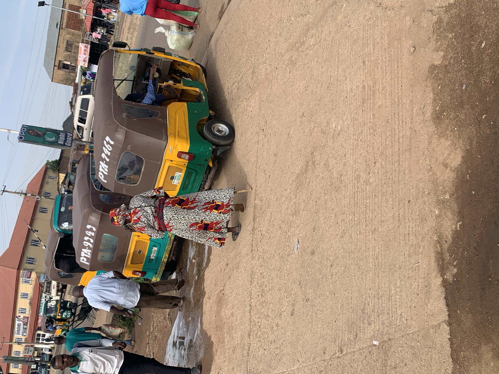 The image depicts a street scene in Gura Top, Nigeria, featuring multiple auto-rickshaws, commonly known as 'keke'. In the foreground, a woman wearing a colorful, patterned dress and headscarf walks with the aid of a stick. Behind her, two auto-rickshaws are parked side-by-side. The one on the left has the registration number PTA-9343 visible on its side, and the one on the right shows PTA-7467. Both vehicles are yellow and green. Several individuals are present in the mid-ground; a man in a white t-shirt and dark pants walks towards the left, and another man in a light blue shirt and brown trousers stands near the auto-rickshaws. In the background, buildings line the street, including one with signage that appears to indicate a pharmacy. The ground is paved and appears somewhat worn, with some wet patches. The sky is overcast.
