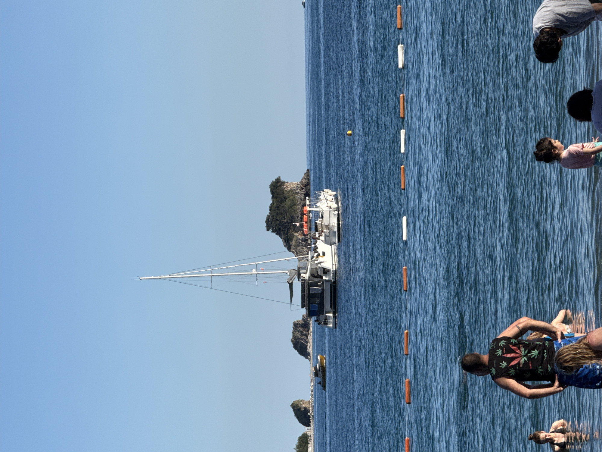 This image depicts a sunny coastal scene in Budva, Montenegro, characterized by a clear blue sky, calm blue sea, and distant rocky islands. The upper third of the image is filled by a uniform, cloudless light blue sky. Below, the deep blue sea occupies the majority of the frame, showing gentle ripples on its surface. Across the mid-ground, a horizontal line of six cylindrical buoys, alternating between orange and white, demarcates an area of the water. A single spherical yellow buoy floats to the right of the primary vessel. Several landmasses are visible on the horizon. On the far left, a light-colored beach or small land area with sparse vegetation and a small white structure extends towards a rocky outcrop. Moving right, multiple dark, rocky islands topped with green foliage emerge from the water. The largest of these islands on the right features dense greenery and a cliff-like structure. Three distinct vessels are present in the mid-ground: 1. A large, white multi-hull sailing yacht (likely a catamaran or trimaran) is positioned centrally. It features a tall, slender white mast with rigging lines, a dark-colored superstructure with multiple windows, and a partially deployed or covered sail. A smaller white motorboat is attached to its stern. 2. A yellow-hulled motorboat with a dark cabin is navigating to the left, situated between the large sailing yacht and the leftmost rocky island. 3. A white motorboat with an orange canopy is partially visible on the right, positioned behind the large sailing yacht and near the large rocky island. A very small, distant boat is faintly discernible on the far right horizon. In the foreground, several individuals are wading or standing in the shallow sea water: * A man, viewed from the back, stands centrally, wearing a black sleeveless shirt adorned with multiple red and green cannabis leaf designs. He has short dark hair, blue swim shorts, and a tattoo on his right upper arm. * To his left, a woman with dark hair tied back is partially submerged to her chest, wearing a dark bikini top, and looking towards the left. * Directly in front of the central man, a person with light brown or blonde hair is partially visible, appearing to be bending or kneeling in the water. * To the right, a small child with dark hair, wearing a pink top and light blue bottoms, stands in the water, looking downwards. An