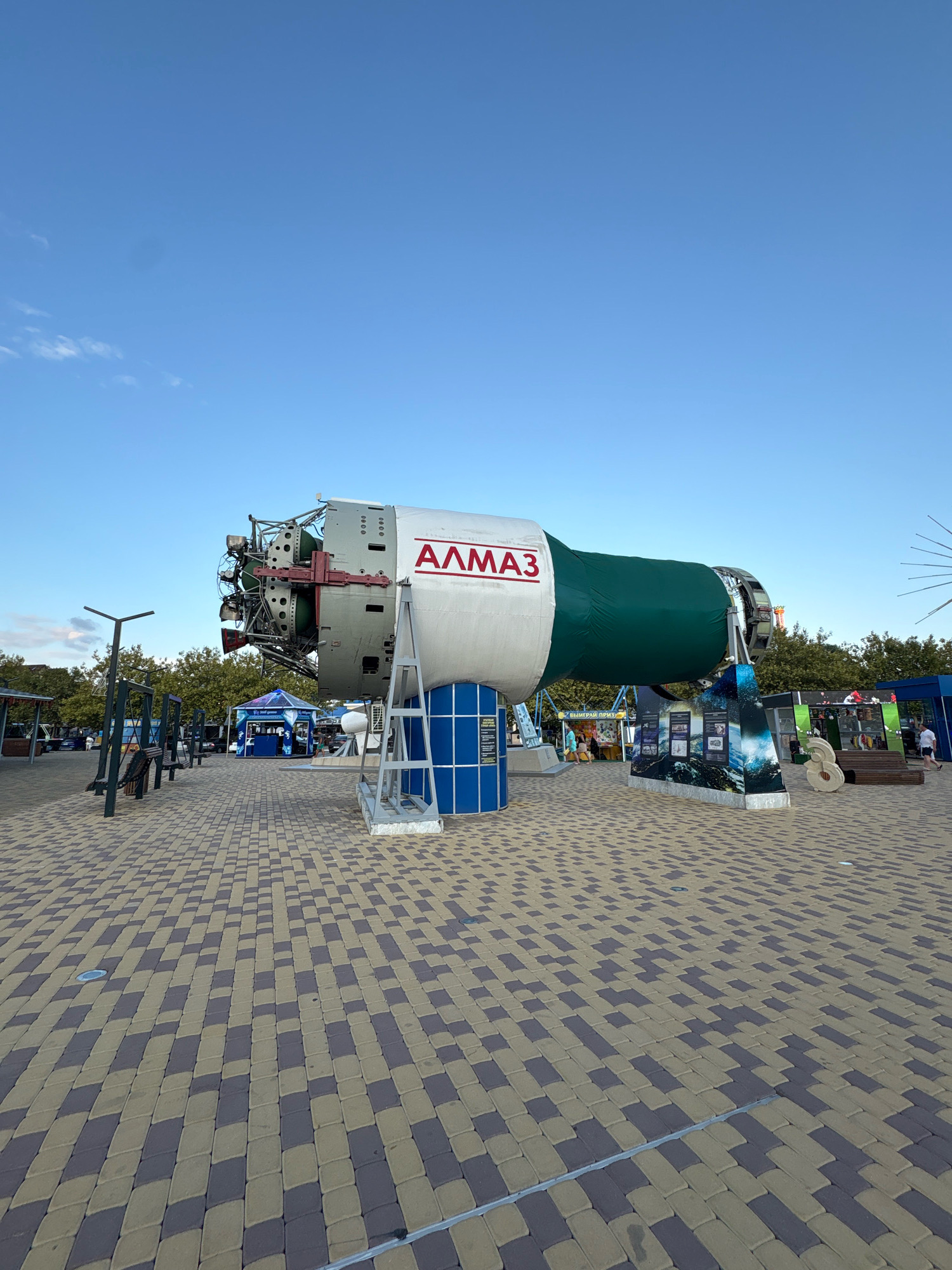 An outdoor, ground-level, full-frame, eye-level shot features a large, white and green cylindrical object, identified by red Cyrillic lettering as "АЛМАЗ" (ALMAZ), on display in a public area. The object, appearing to be a model of a spacecraft or rocket, is supported by a metal framework. The surrounding area is paved with a pattern of tan and dark gray rectangular bricks. In the background, trees and various structures are visible, including tents, buildings with signage, and a few people. The sky above is clear and blue. The scene is located in Arkhipo-Osipovka, Russia, specifically at the "Dubki" park, which serves as an open-air museum dedicated to space exploration.