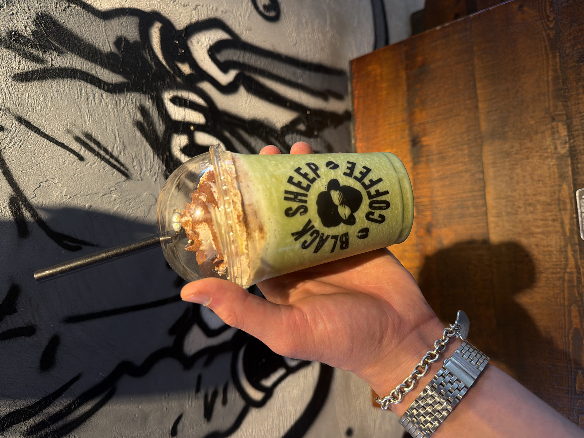 A close-up, high-angle shot shows a person's left hand holding a clear plastic cup containing a green blended beverage, likely a matcha latte, with whipped cream and chocolate powder on top. The cup has a black logo that reads "BLACK SHEEP COFFEE." A black straw extends from the cup. The person is wearing a silver bracelet on their wrist. The background is a textured grey wall with black graffiti. The scene is lit by overhead lighting, casting shadows. The lower portion of the image shows a wooden table surface. The image's composition and the "Black Sheep Coffee" branding suggest it was taken at a Black Sheep Coffee location, a coffee shop chain with multiple branches in London, United Kingdom.