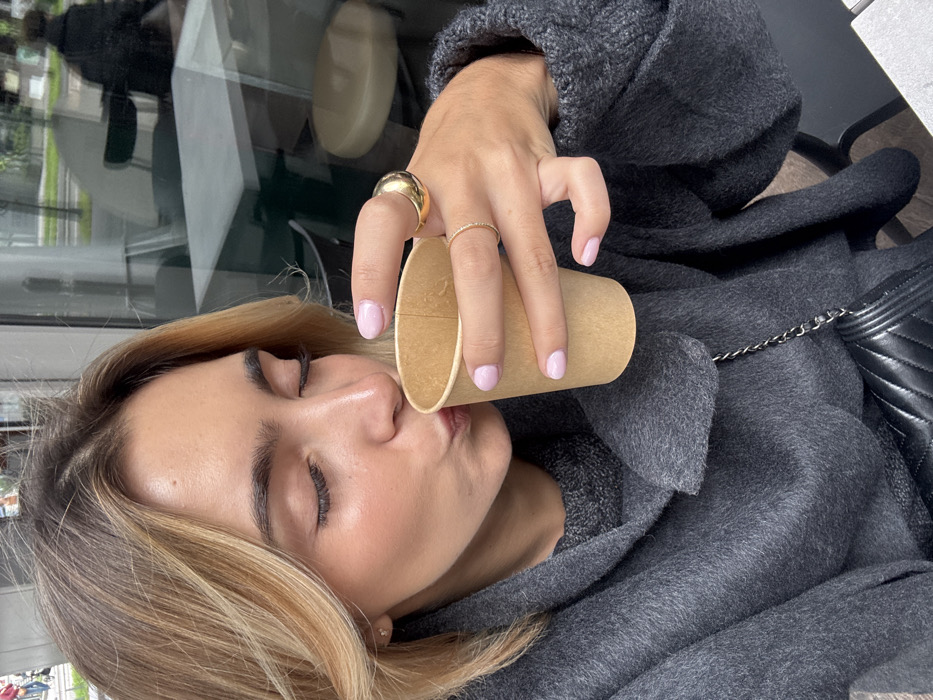 A young adult female is depicted in a close-up, chest-up shot, actively drinking from a plain, light brown paper cup. Her eyes are closed, and her lips are pursed towards the cup's rim. She possesses light brown hair with blonde highlights and a visible small gold stud earring in her left earlobe. Her right hand, featuring light pink manicured fingernails, holds the cup; a large, smooth gold dome ring is present on her right index finger, and a thin gold band ring on her right ring finger. She is dressed in a dark gray, textured sweater or coat, with a darker gray knit top visible beneath the wide collar. A black quilted leather handbag with a silver chain strap is slung over her left shoulder, resting on her lap. The background is a blurred reflection from a large glass window, indicating an outdoor urban environment beyond, with visible elements including green foliage, a light-colored building, and a dark, indistinct human silhouette walking leftward. Interior reflections show parts of minimalist white-topped tables and dark chairs, suggesting an indoor cafe or restaurant setting in Vaulx-en-Velin, France.