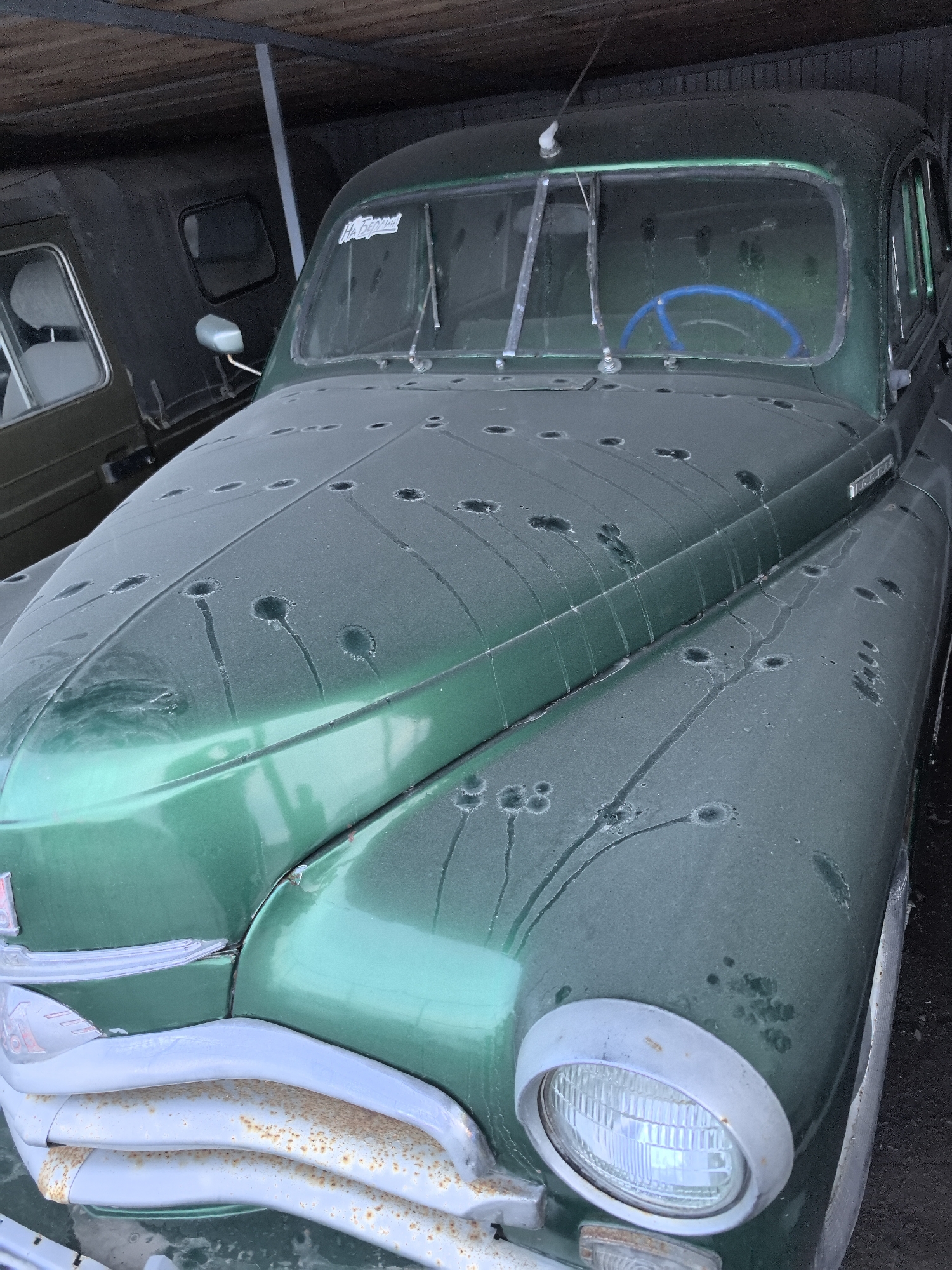 The image shows a green, heavily weathered GAZ-M20 Pobeda car in what appears to be a garage or storage area. The car is covered in dust or a similar particulate, creating numerous circular patterns on the hood and front fenders, likely caused by moisture accumulating on the dusty surface. The front bumper is corroded. The interior is visible through the windshield, with a blue steering wheel. A dark-colored vehicle with a boxy shape is parked to the left of the Pobeda. The location is Bol'shoi Mashlyak, Russia.