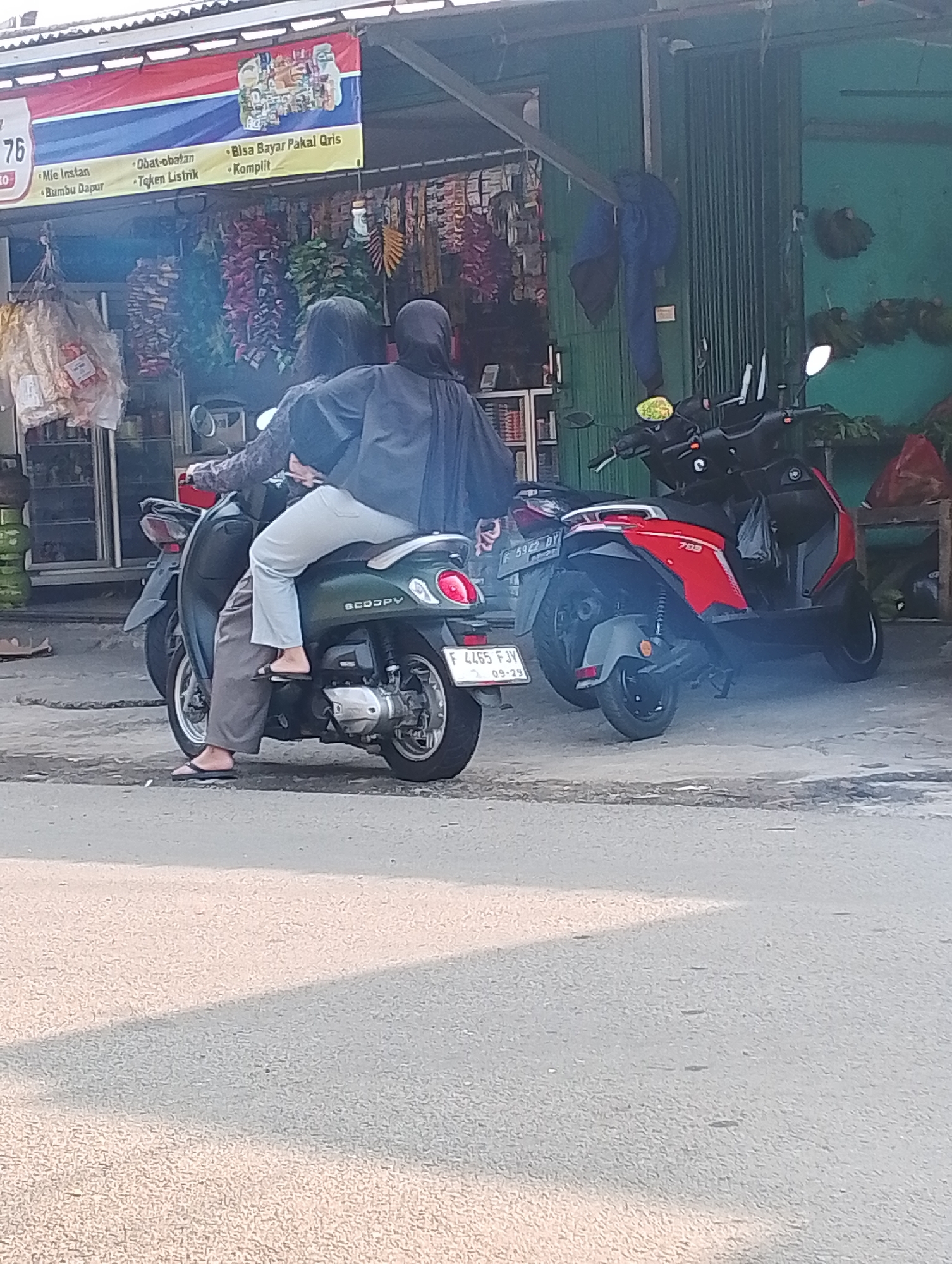 Two individuals are seated on a dark green scooter in front of a storefront in Bogor, Indonesia. The person in the back is wearing a dark headscarf and dark clothing. The person in the front is wearing light blue pants and a patterned long-sleeved shirt. The scooter has the brand name "SCOOPY" visible on its side and a license plate reading "F 4465 FJV" with a sticker below it. Another red scooter is parked behind the green one. The storefront appears to be a small shop selling various goods, with bags of products and hanging items visible. The ground is paved with asphalt.