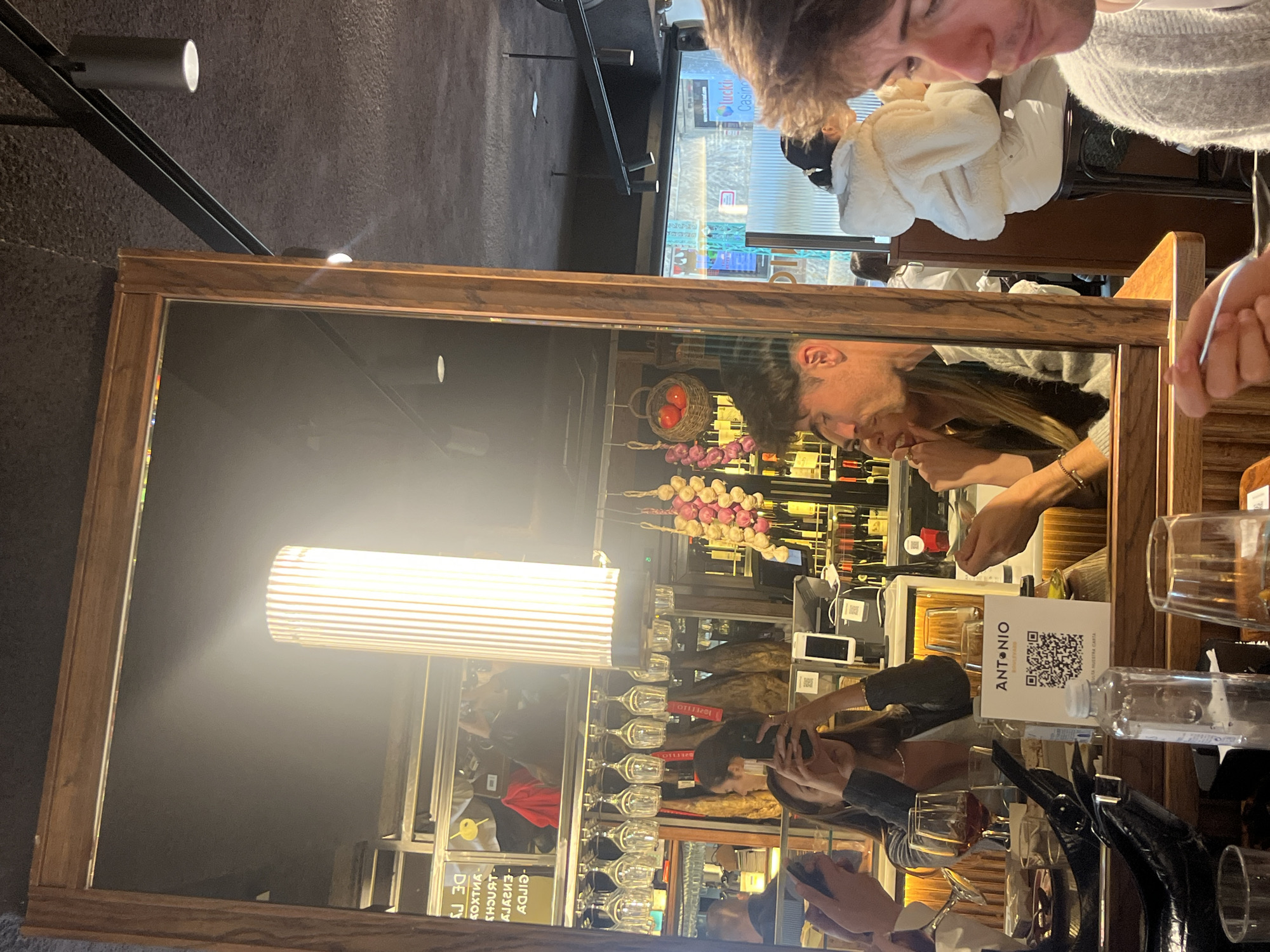 The image shows the reflection of a restaurant scene in a large, wooden-framed mirror. The mirror reflects a well-lit bar area with wine glasses hanging above the counter. A sign that reads "Antonio" is visible. People are sitting at the bar, one appearing to use their phone. A basket of tomatoes and strings of garlic hang behind the bar.
The actual foreground of the image shows people sitting at a table, with a man in a grey sweater visible. The location is presumed to be in San Sebastián, Spain, based on the name of the restaurant.