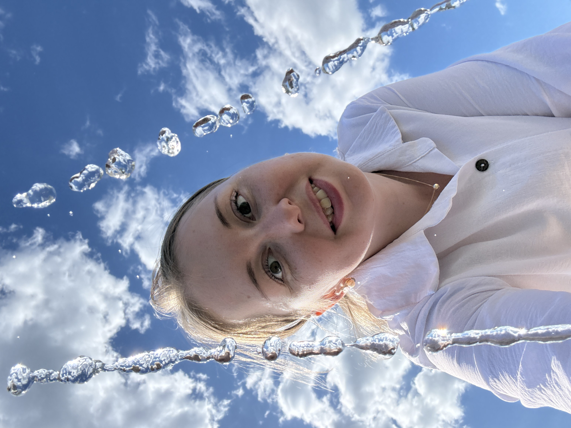 A young Caucasian woman is depicted in a low-angle, close-up outdoor shot, looking directly at the camera with a slight smile. Her blonde hair is pulled back, with sunlit wisps framing her face. She wears a white collared shirt with long sleeves; a single dark button is visible on the placket, and a delicate gold chain necklace with a small spherical pendant rests on her chest. A light-colored drop earring is visible on her right ear.
Streams and individual droplets of water are falling from above, framing her on both sides. On the left, a continuous, segmented column of water appears to be falling, reflecting bright sunlight. On the right, several distinct, spherical water droplets are captured mid-air. The background is a bright blue sky dotted with scattered white cumulus clouds, all illuminated by strong natural daylight, suggesting a sunny outdoor setting in the city of Unknown, Unknown.